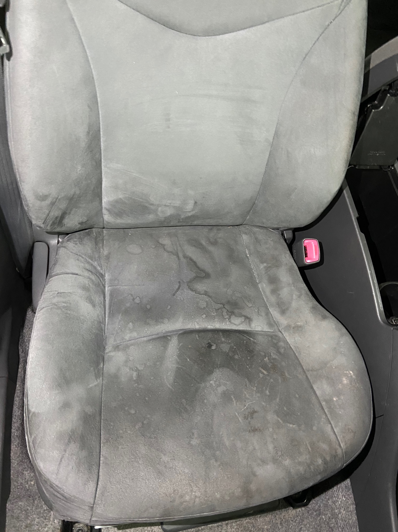 Szare, mocno zabrudzone welurowe siedzenie samochodowe z widocznymi plamami i zaciekami, różowy zatrzask pasa bezpieczeństwa