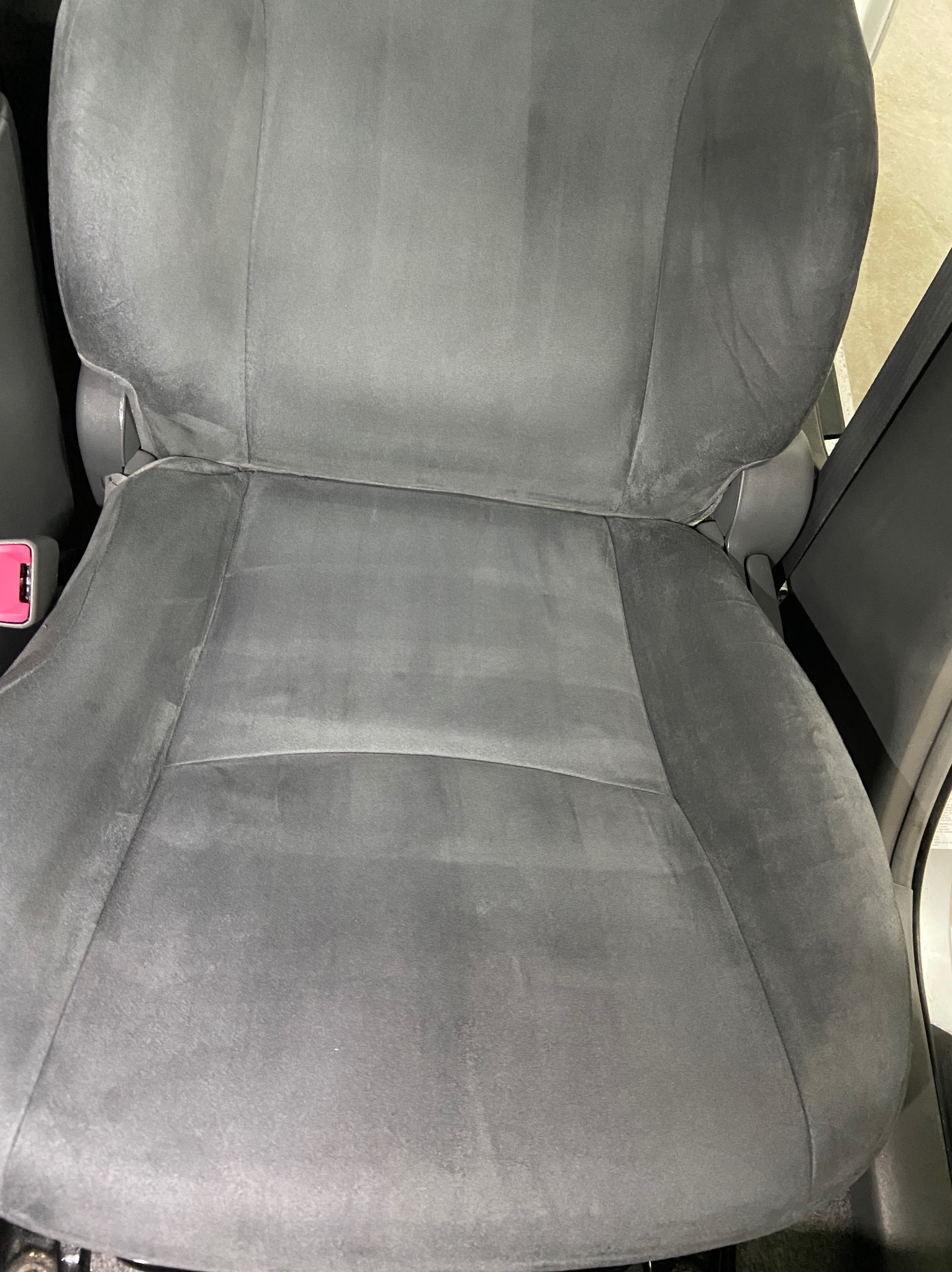 Szare welurowe siedzenie samochodowe po praniu tapicerki, widoczne ślady po czyszczeniu, zapięcie pasów bezpieczeństwa w kolorze różowym.