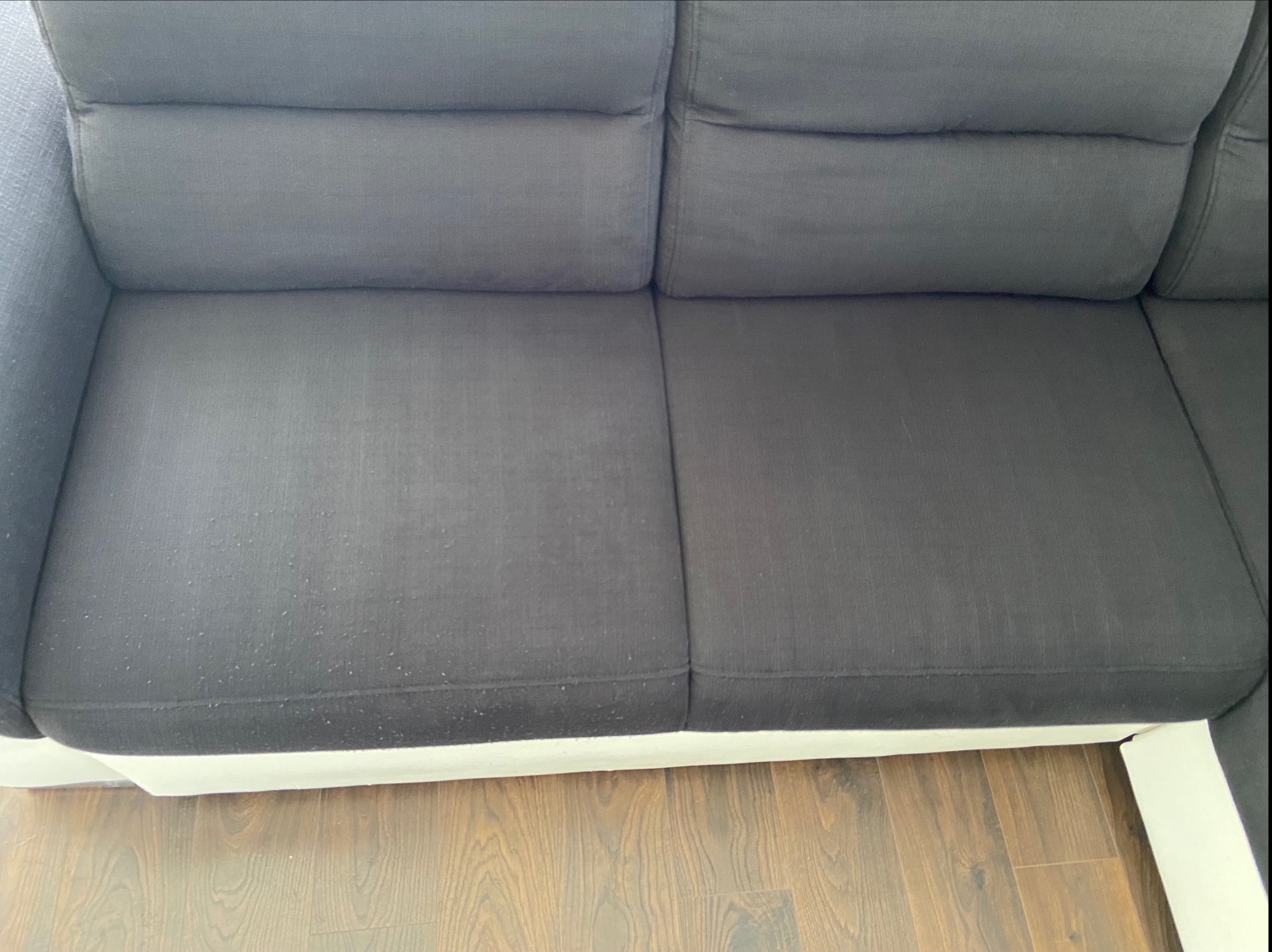 Szara sofa z widocznymi zabrudzeniami i plamkami na tapicerce, biała podstawa, drewniana podłoga.