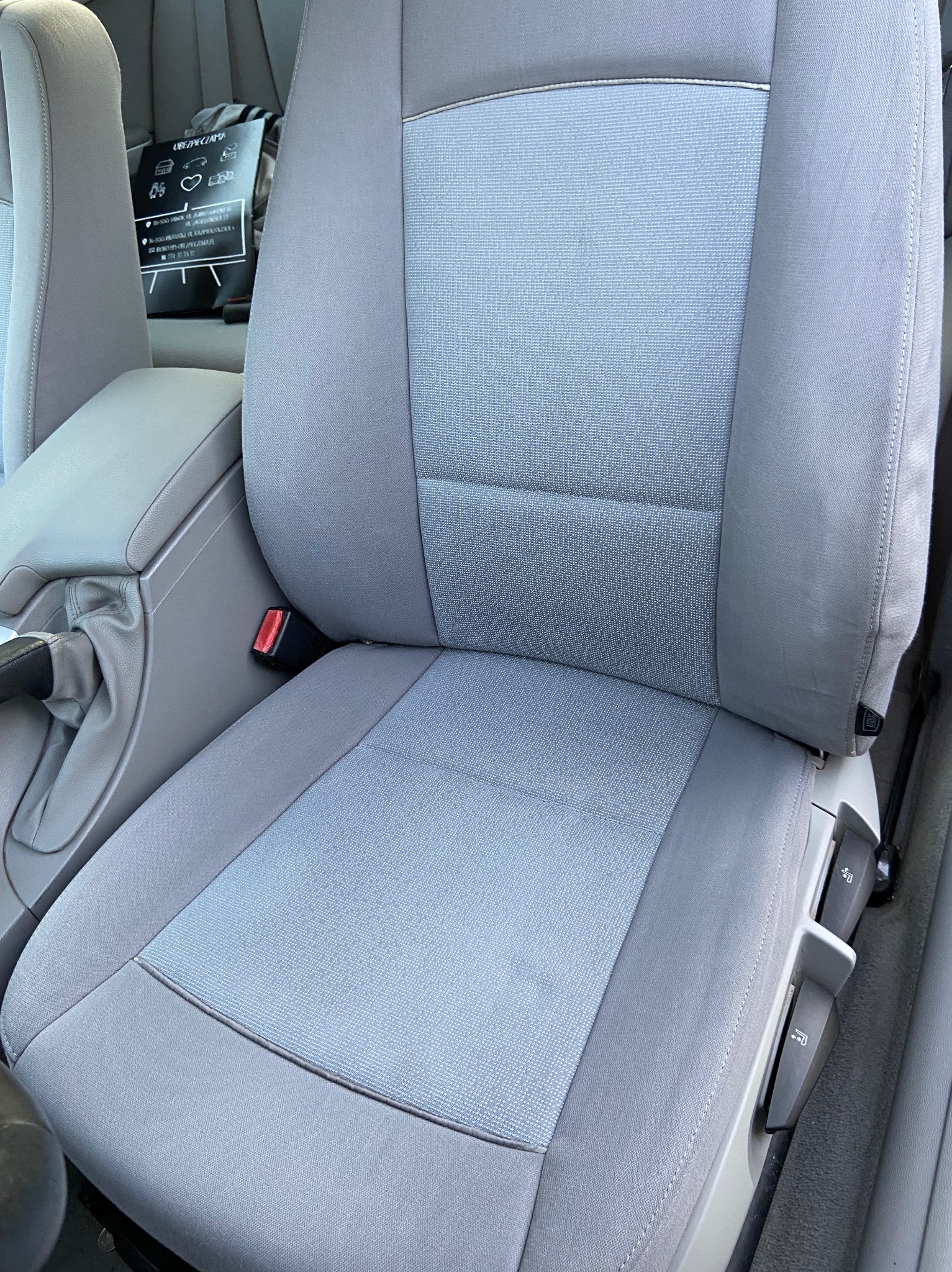 Szare, materiałowe siedzenie samochodowe z widocznym pasem bezpieczeństwa i regulacją. Na zagłówku widoczny czarny worek z instrukcją.