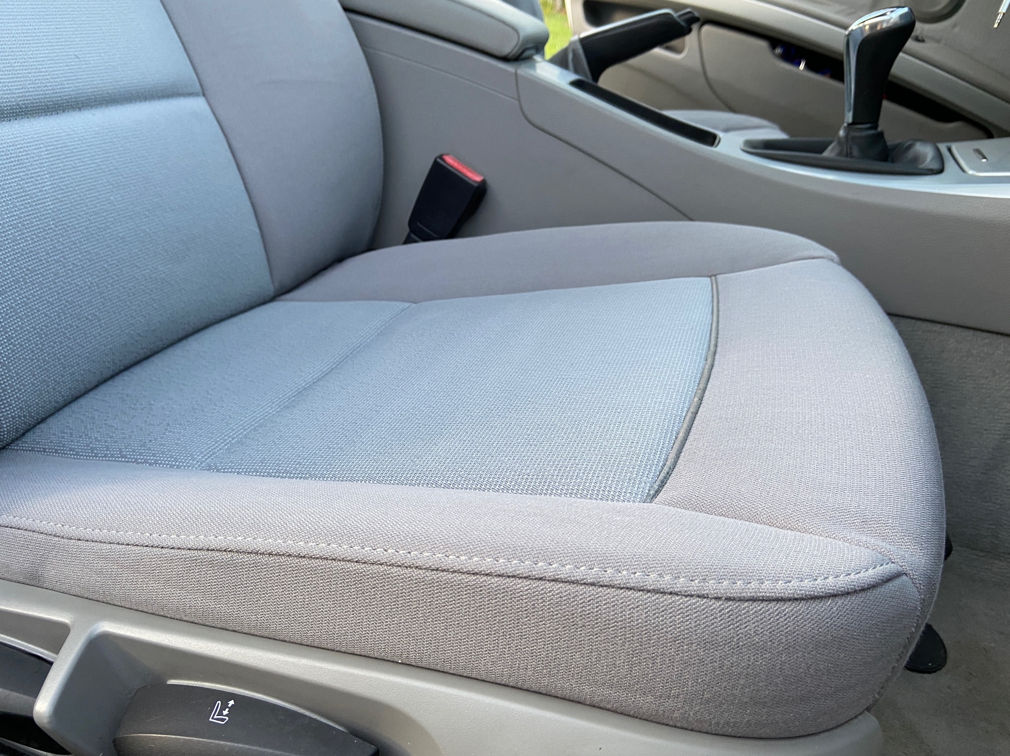 Szare, materiałowe siedzenie samochodowe z widocznym pasem bezpieczeństwa, dźwignią zmiany biegów i elementami regulacji fotela.