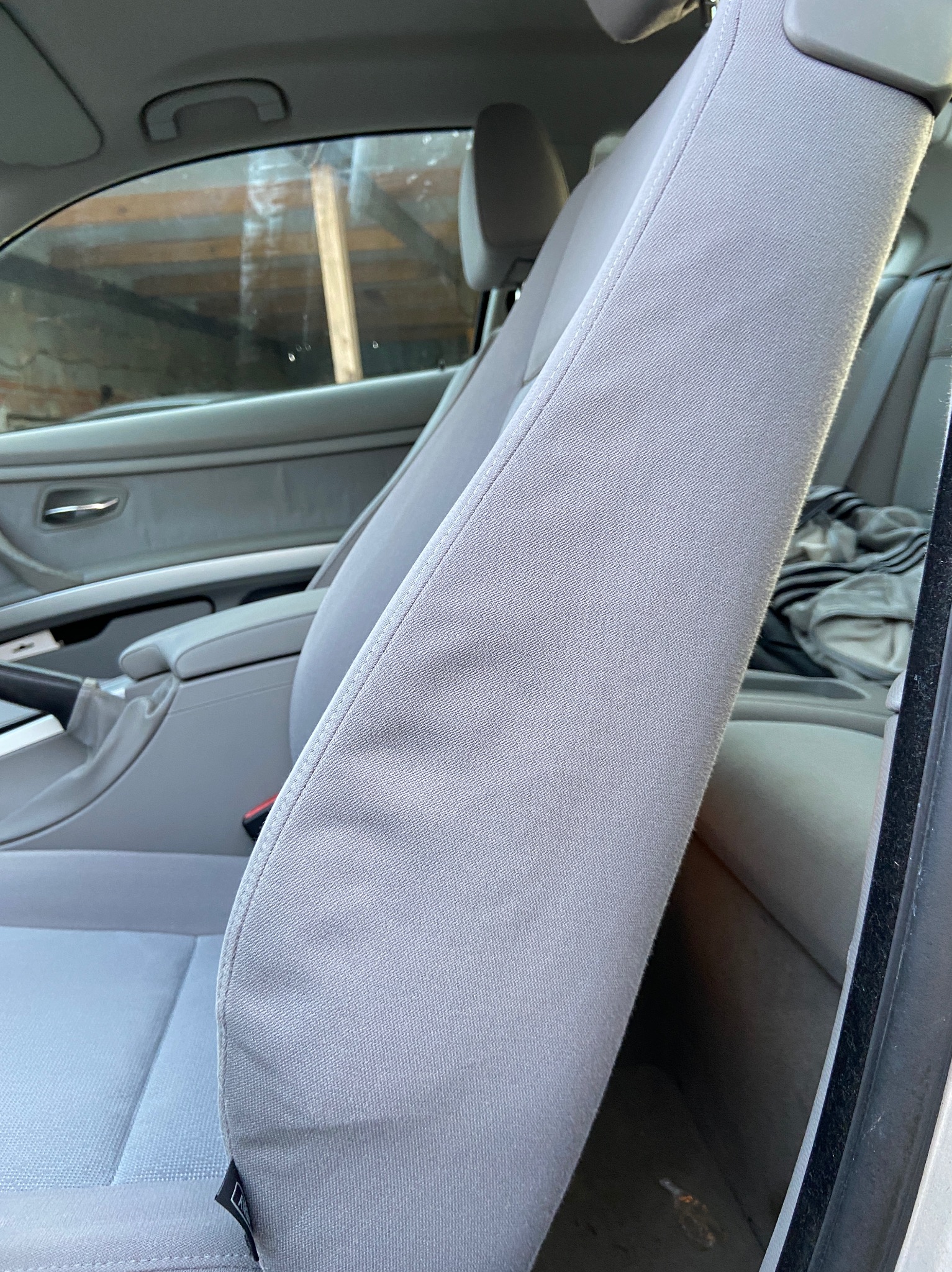 Jasnoszare, materiałowe siedzenie samochodowe, widoczny zagłówek, podłokietnik i fragment drzwi. W tle niedbale rzucone ubrania na tylnym siedzeniu.
