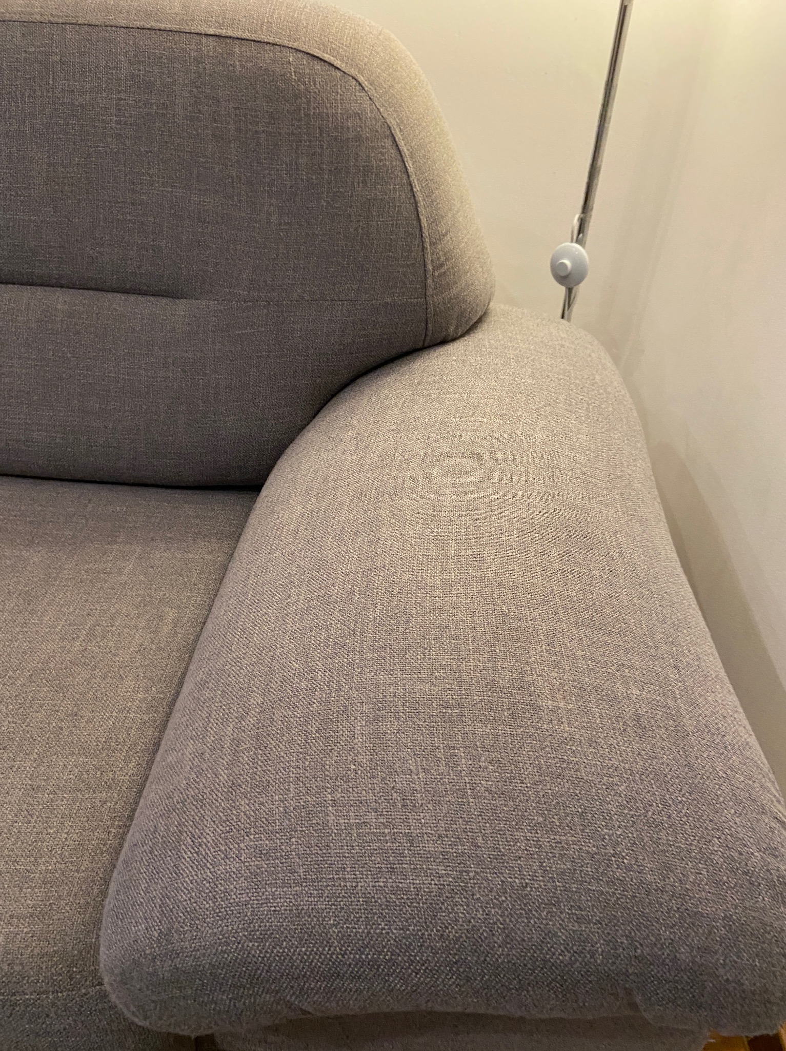 Fragment szarej, tapicerowanej kanapy z widocznym podłokietnikiem i oparciem, oświetlony lampą stojącą w tle.