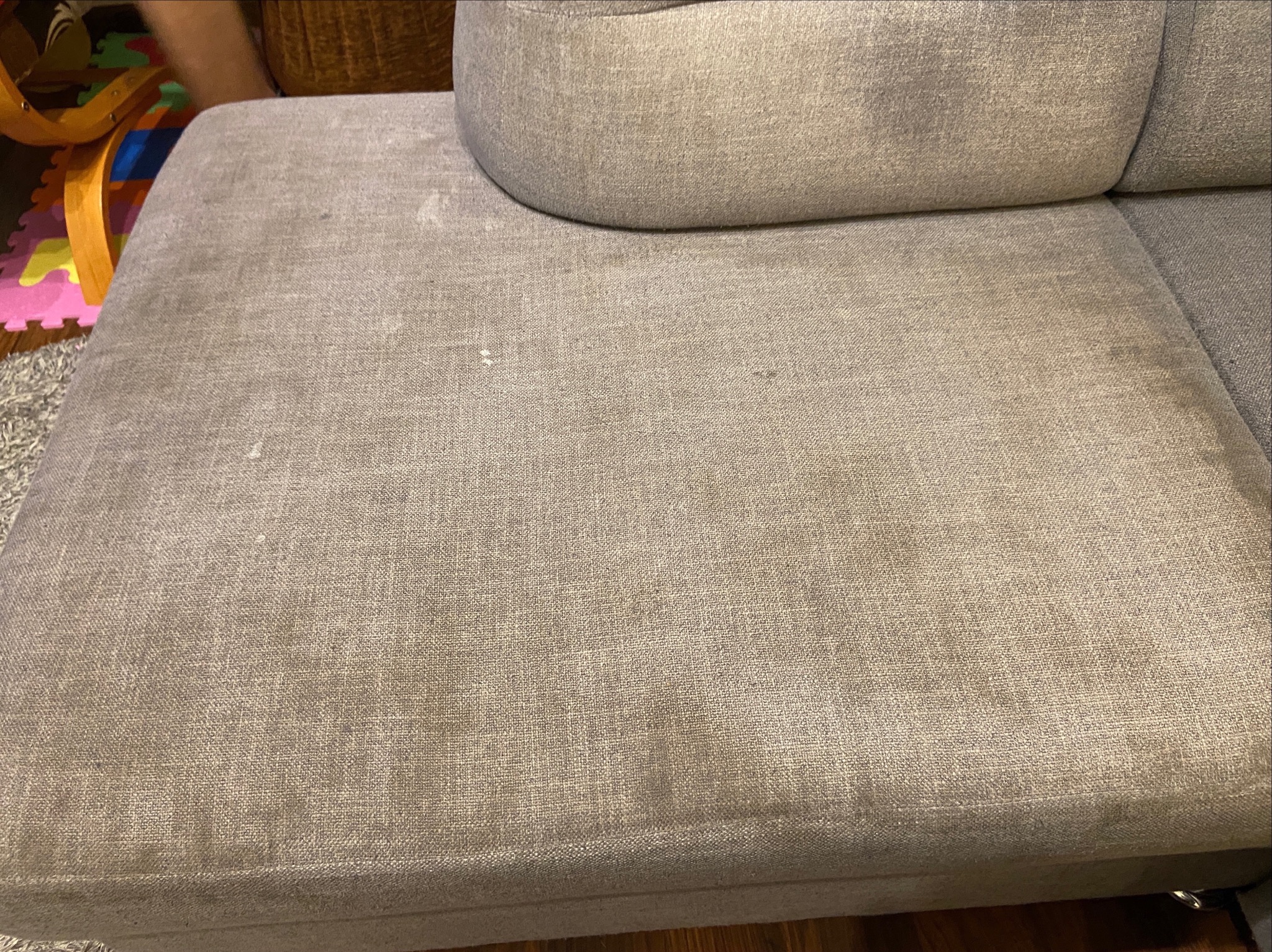 Fragment sofy z beżową tapicerką, widoczne plamy na siedzisku, w tle zabawki dziecięce.