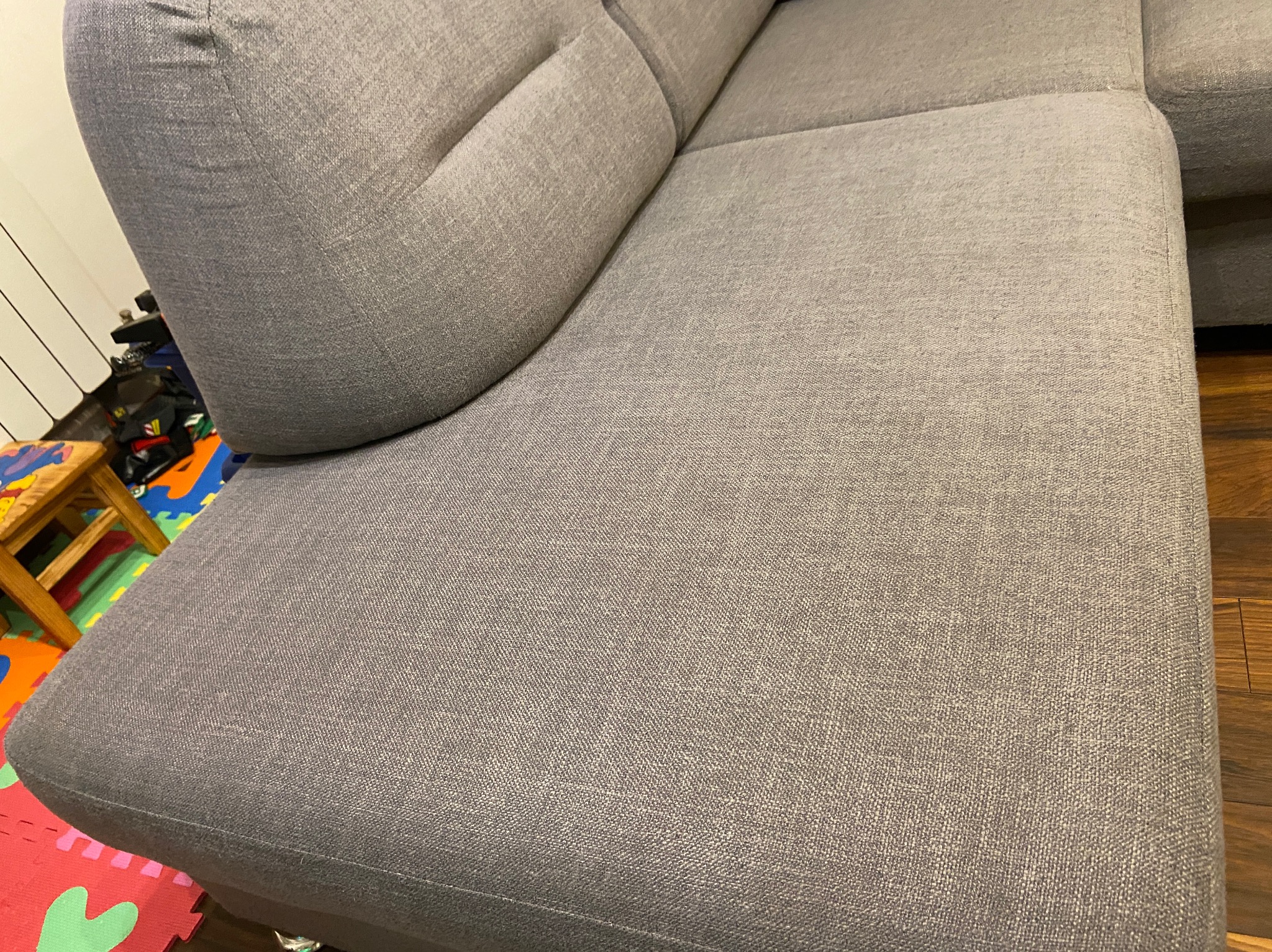 Fragment szarej, narożnej sofy z widoczną strukturą tkaniny, na podłodze panele oraz dziecięca mata piankowa.
