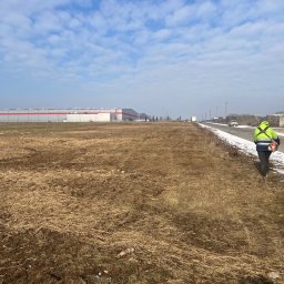 Kruszer-Warka Wioleta Gwiazda - Pracownik w jaskrawej kurtce z kosiarką spalinową na tle pola i budynku magazynowego. Pejzaż z resztkami śniegu i błękitnym niebem.