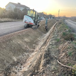 Kruszer-Warka Wioleta Gwiazda - Mini koparka Bobcat 323 tworzy rów wzdłuż drogi gruntowej oświetlonej zachodzącym słońcem, z budynkiem w tle. Widoczne świeże ślady pracy.