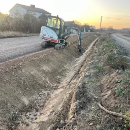 Mini koparka Bobcat 323 tworzy rów wzdłuż drogi gruntowej oświetlonej zachodzącym słońcem, z budynkiem w tle. Widoczne świeże ślady pracy.