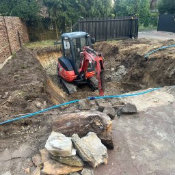 Kruszer-Warka Wioleta Gwiazda - Czerwony mini-koparka Kubota w wykopie, widoczne ślady świeżych prac ziemnych, niebieski wąż, fragmenty kostki brukowej i ogrodzenia w tle.