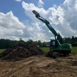 Kruszer-Warka Wioleta Gwiazda - Zielona koparka Komatsu PW170ES z podniesionym ramieniem i łyżką na placu budowy, z hałdą ziemi i śladami opon w tle, pod błękitnym niebem z chmurami.