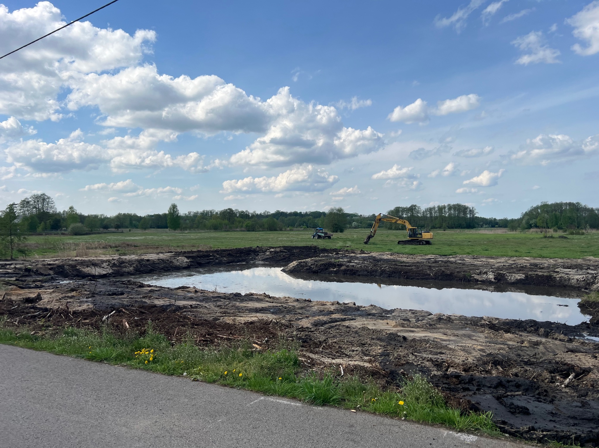 Wykopany staw w trakcie realizacji z użyciem koparki i traktora na tle zielonego krajobrazu pod błękitnym niebem z białymi chmurami. Woda wypełnia częściowo wykop.