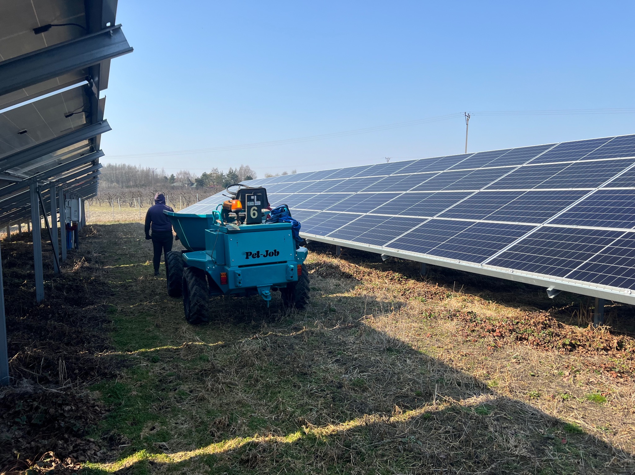 Niebieska mini-wywrotka Pel-Job na trawiastym terenie obok paneli słonecznych, z pracownikiem w tle. Widok z tyłu, słoneczny dzień.