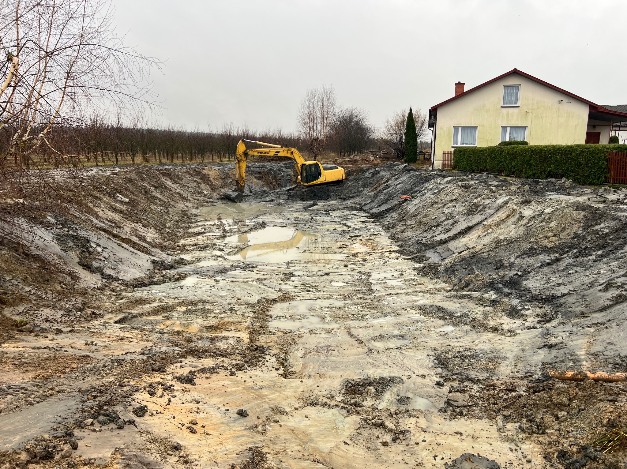 Żółta koparka Komatsu w wykopie pod staw, z błotnistym dnem i kałużami, na tle domu i drzew. Wykopanie stawów.