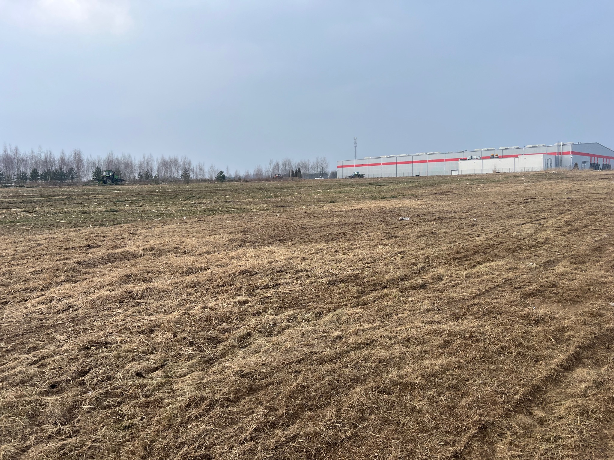 Pole z brązową trawą i budynkiem w tle. Widoczne ślady po pracach ziemnych oraz drzewa. W oddali maszyna budowlana. Pochmurne niebo.