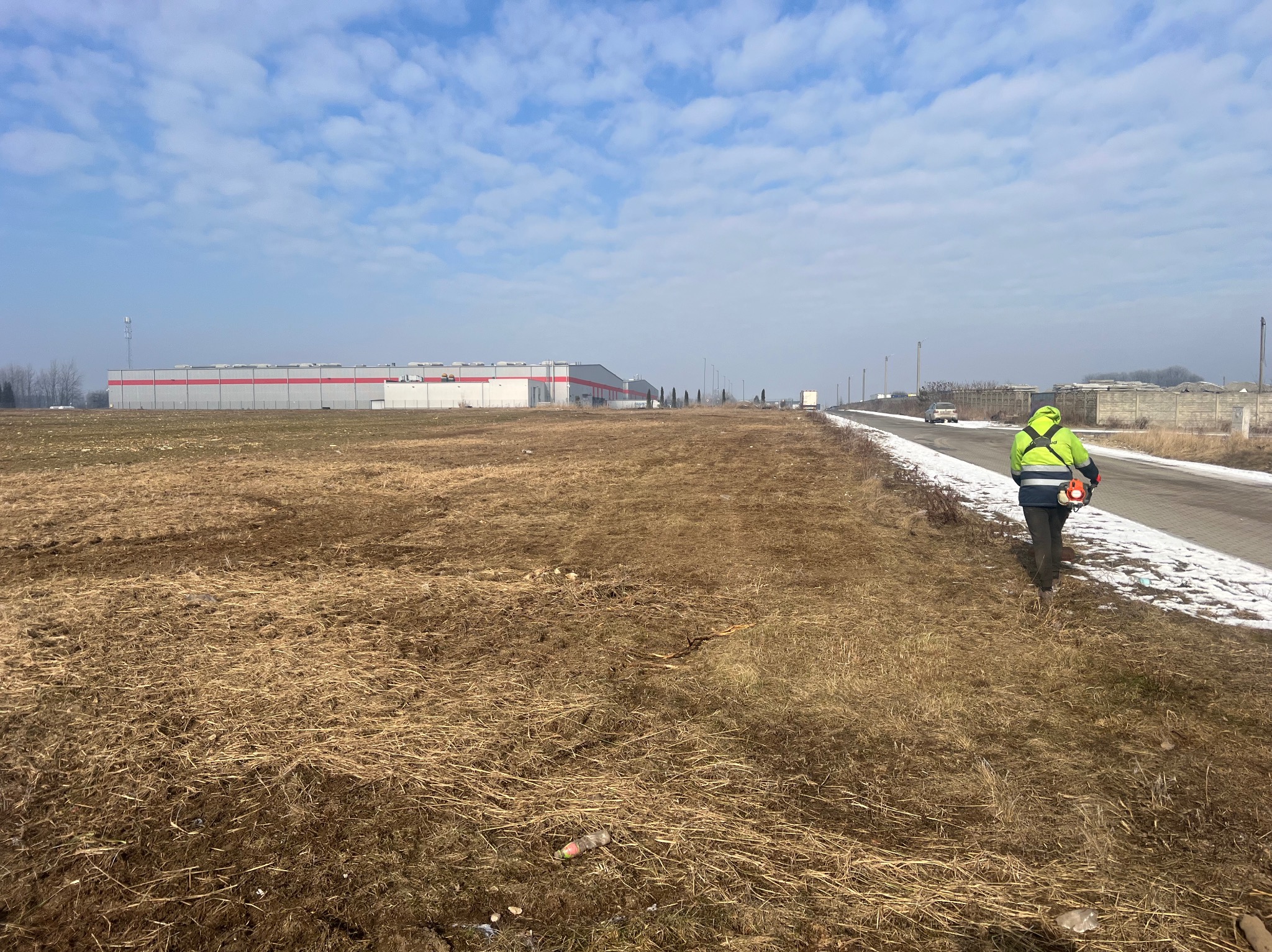 Pracownik w jaskrawej kurtce z kosiarką spalinową na tle pola i budynku magazynowego. Pejzaż z resztkami śniegu i błękitnym niebem.