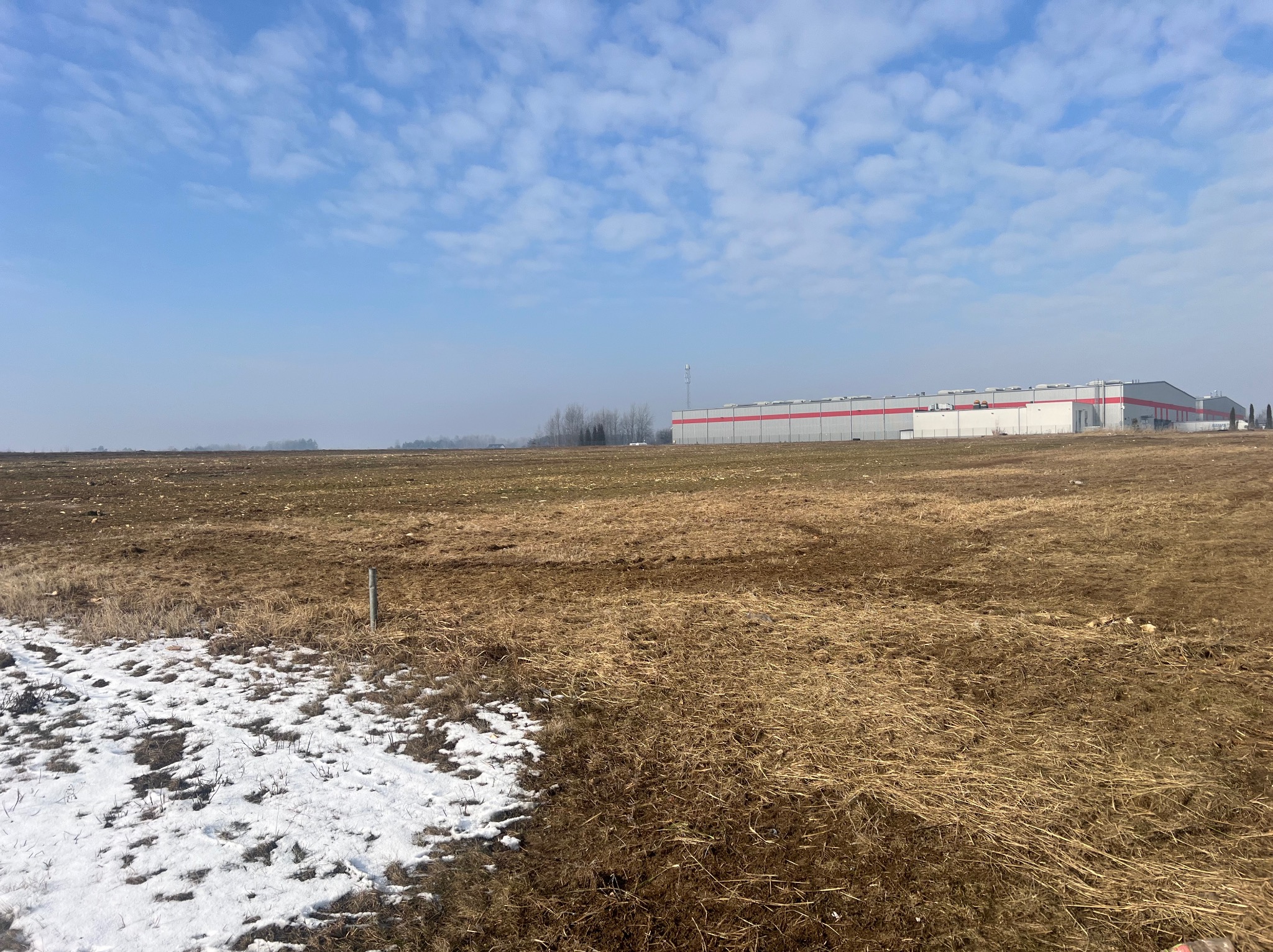 Rozległy teren z częściowo stopniałym śniegiem i budynkiem magazynowym w tle, pod błękitnym niebem z chmurami. Widok na plac budowy lub teren po niwelacji.