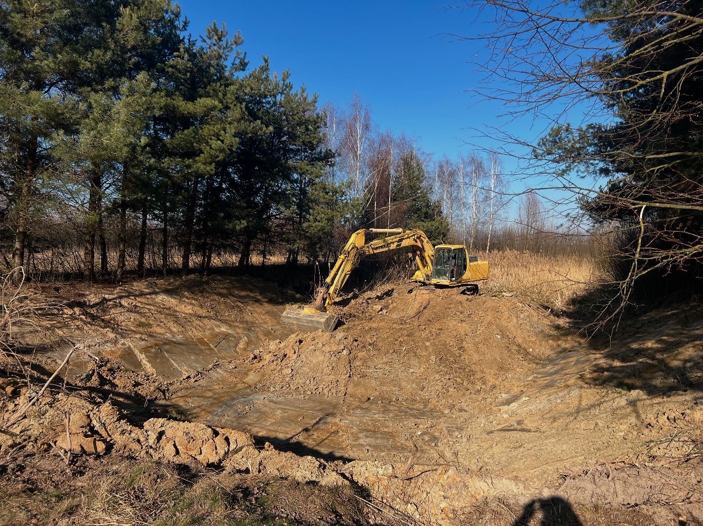 Żółta koparka Komatsu przy wykopanym stawie w otoczeniu drzew iglastych i liściastych pod błękitnym niebem. Widoczny świeży wykop i pryzma ziemi.