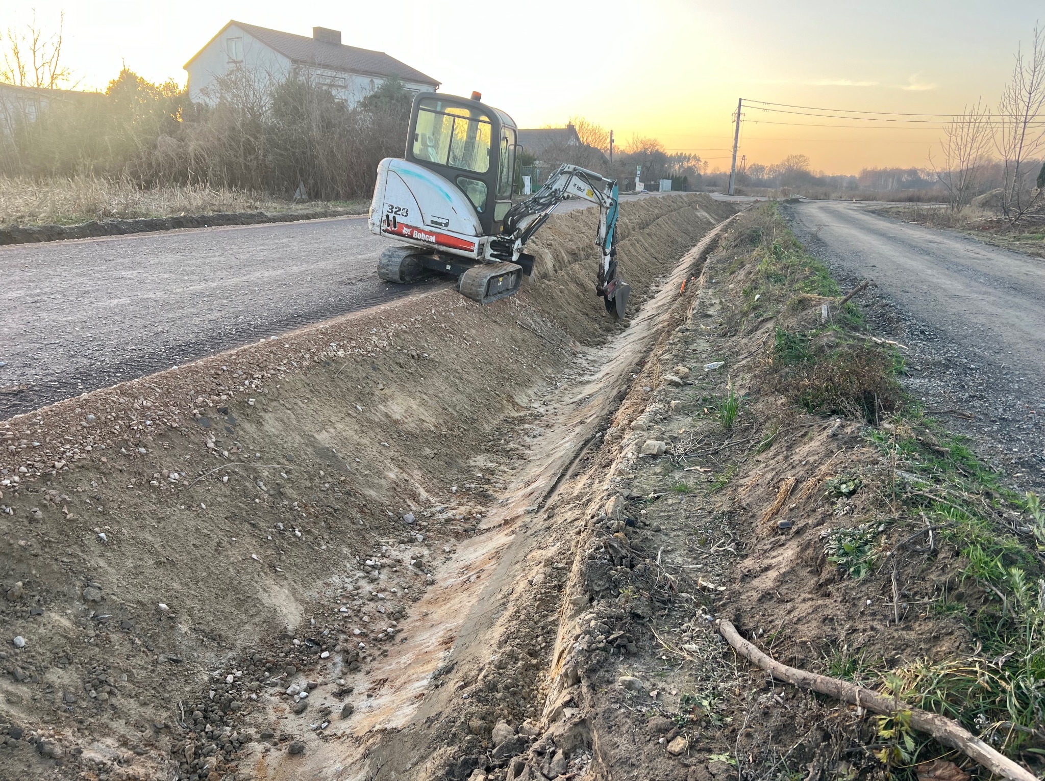 Mini koparka Bobcat 323 tworzy rów wzdłuż drogi gruntowej oświetlonej zachodzącym słońcem, z budynkiem w tle. Widoczne świeże ślady pracy.