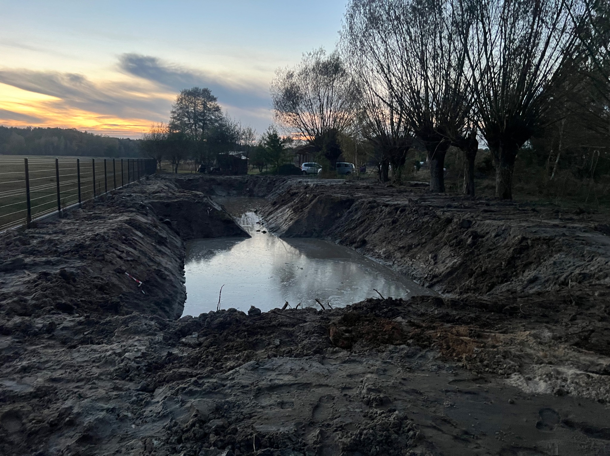 Wykopany staw w ziemi, częściowo wypełniony wodą, otoczony brzegiem z ciemnej ziemi, z widocznymi drzewami i zachodzącym słońcem w tle.