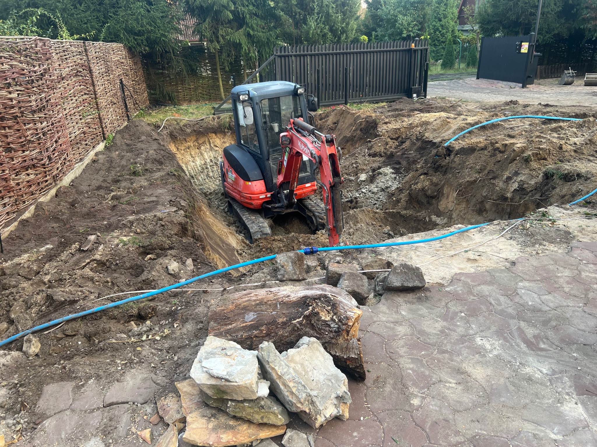 Czerwony mini-koparka Kubota w wykopie, widoczne ślady świeżych prac ziemnych, niebieski wąż, fragmenty kostki brukowej i ogrodzenia w tle.