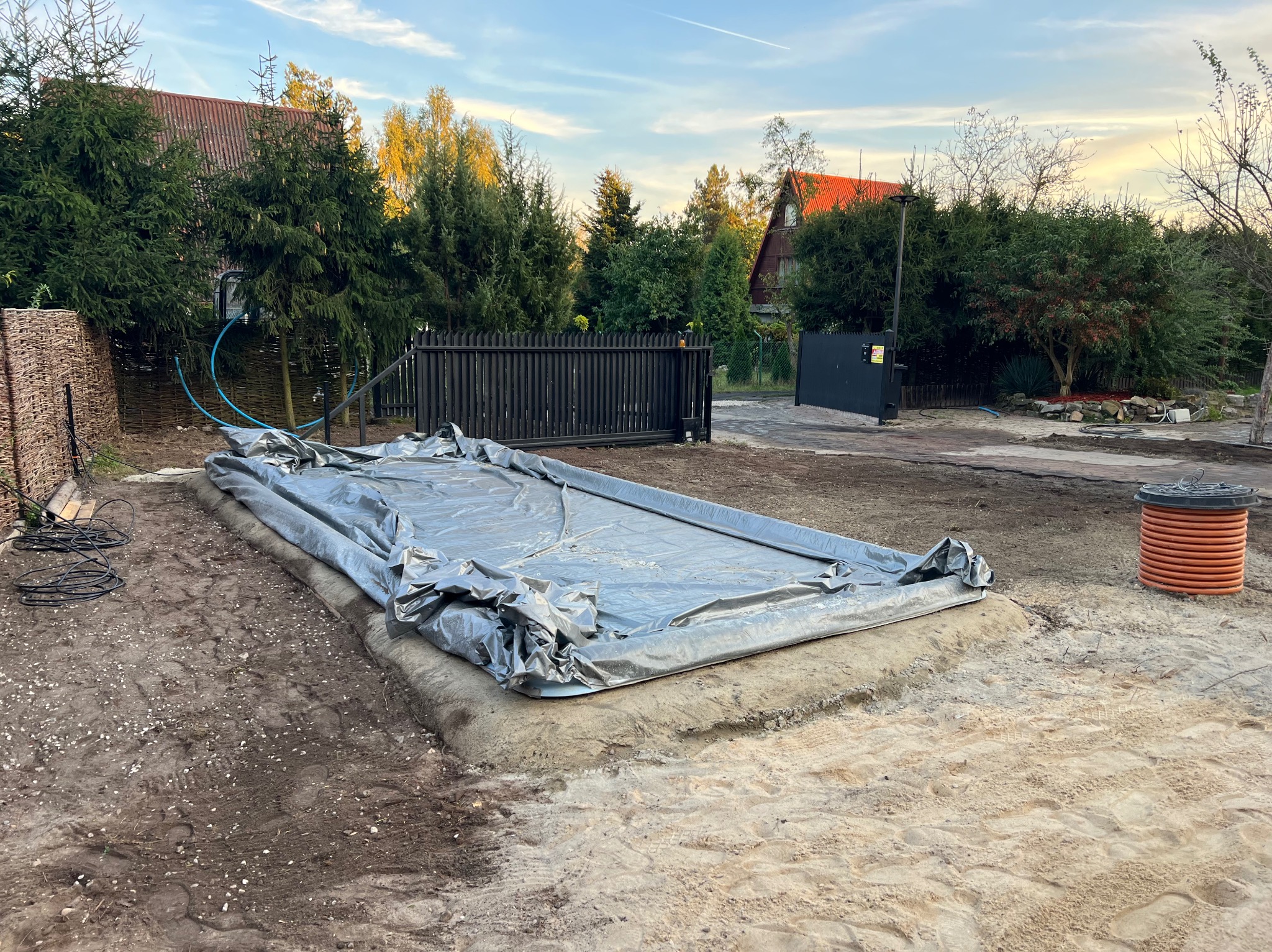 Przygotowany wykop zabezpieczony folią, obok rolka rury drenarskiej. Grunt wokół wyrównany, w tle zieleń i zabudowania.