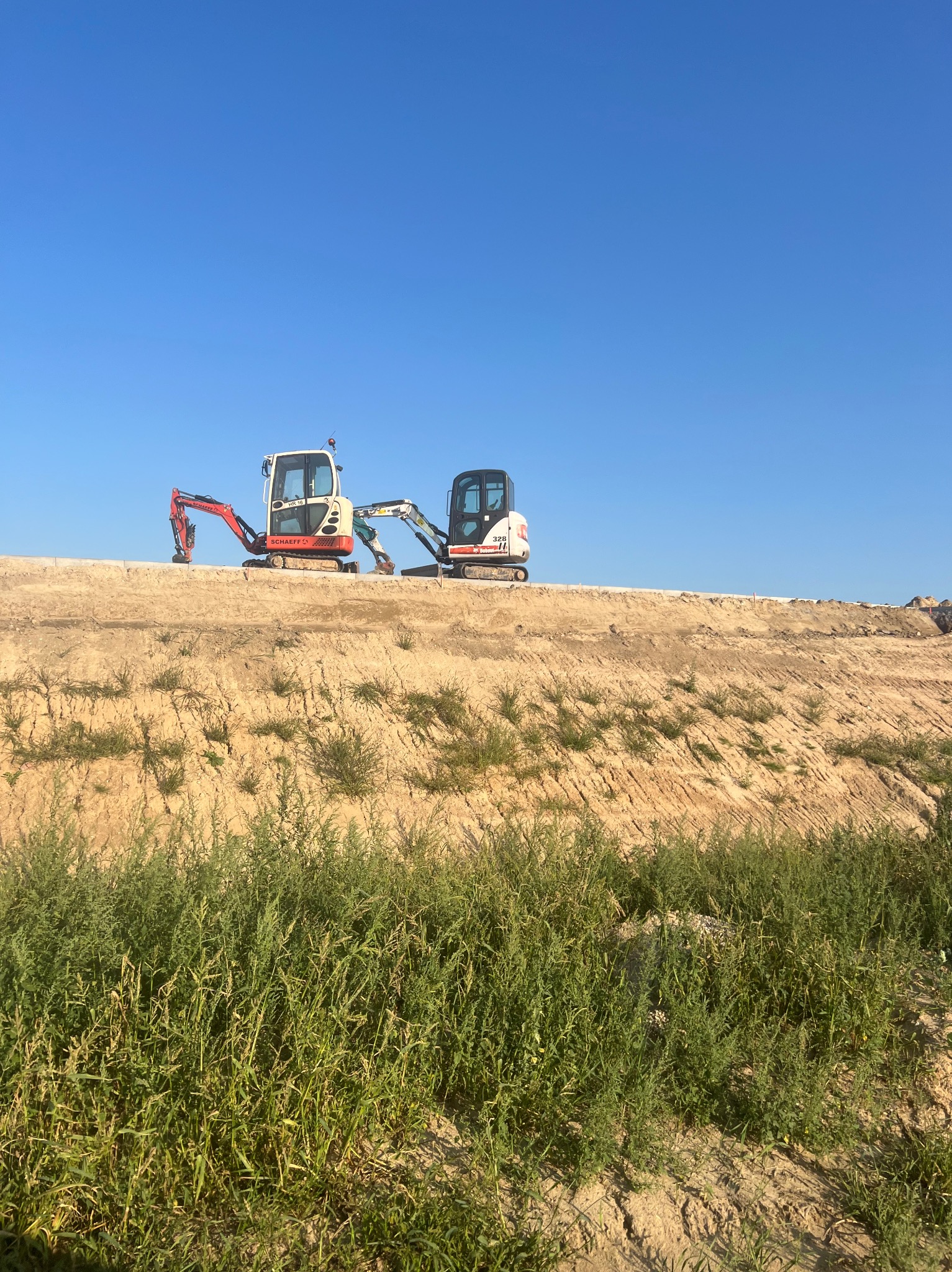 Dwa mini-koparki (Schaeff HR16 i Bobcat 328) na szczycie ziemnego nasypu, na tle błękitnego nieba. Widoczne rośliny na pierwszym planie.