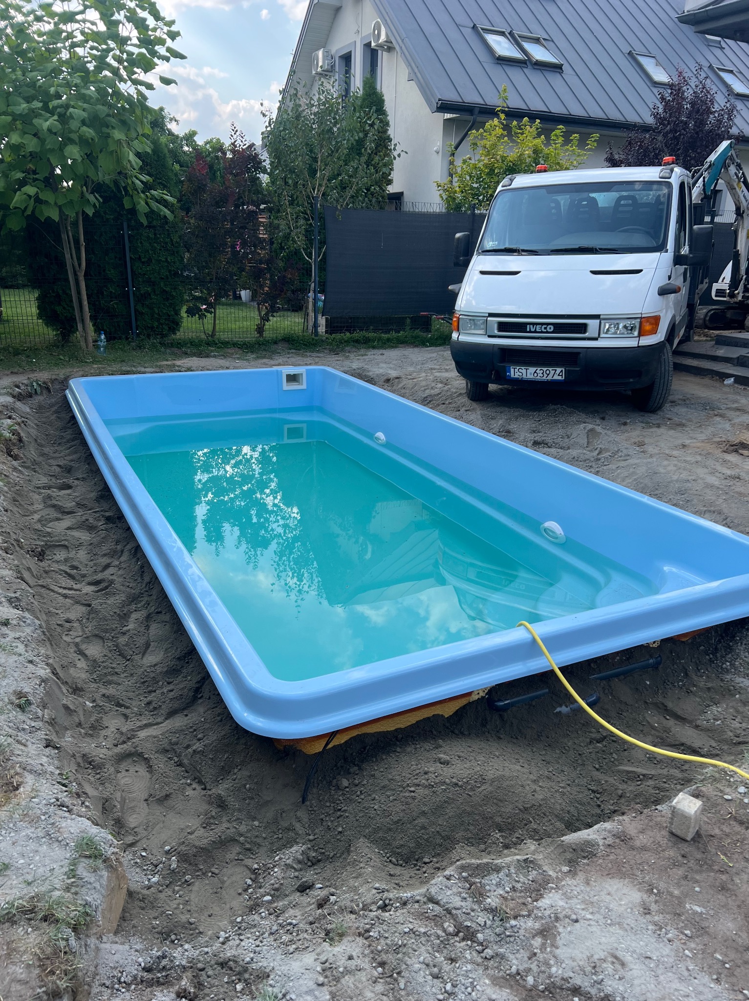 Wkopany basen z błękitną wodą, widoczny biały Iveco i minikoparka w tle. Żółty kabel leży obok basenu. Etap instalacji basenu w ziemi.
