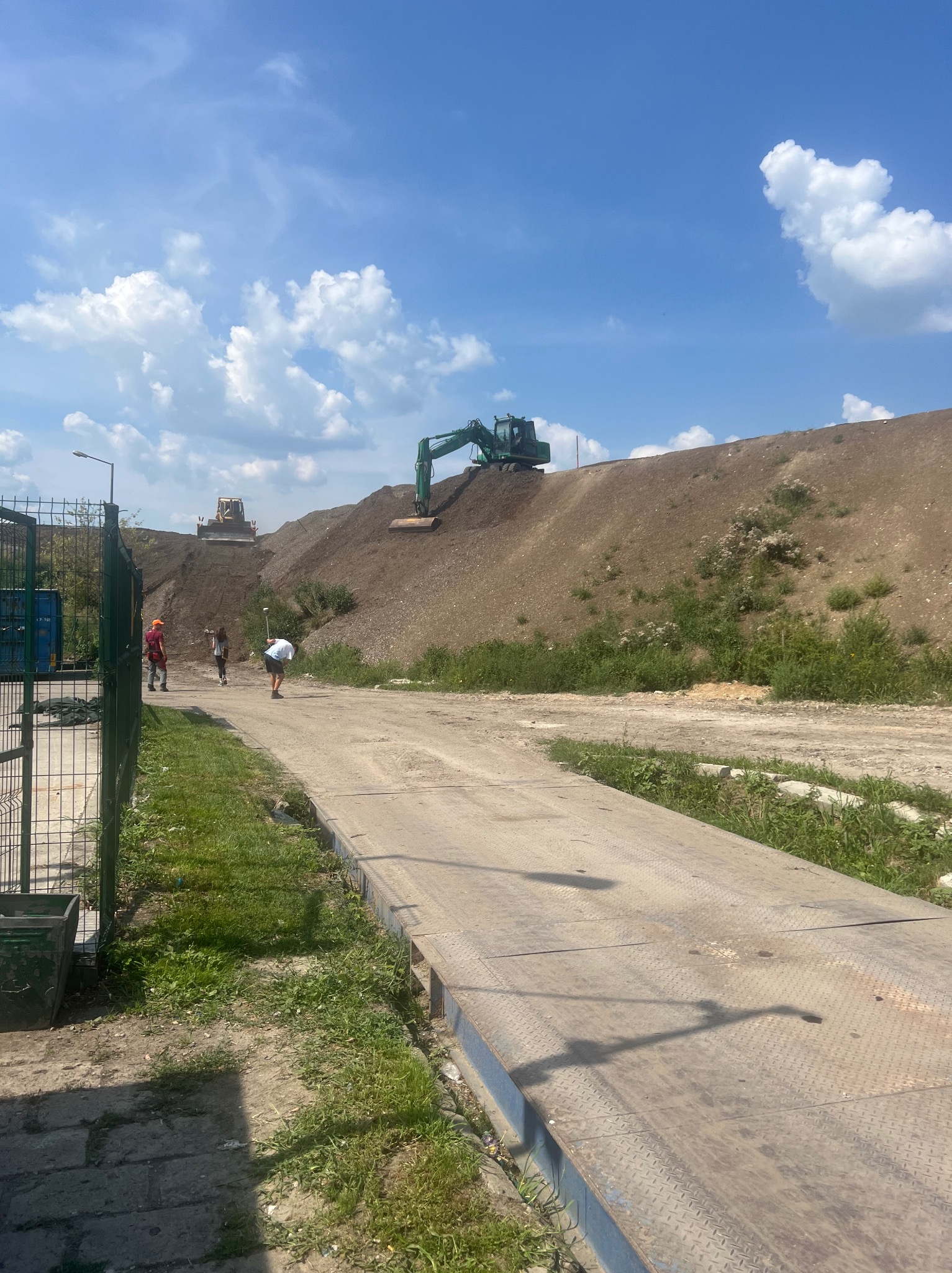 Prace ziemne w toku: koparka na szczycie usypanego wzniesienia, pracownicy w terenie, widoczna waga najazdowa. Błękitne niebo z chmurami w tle.