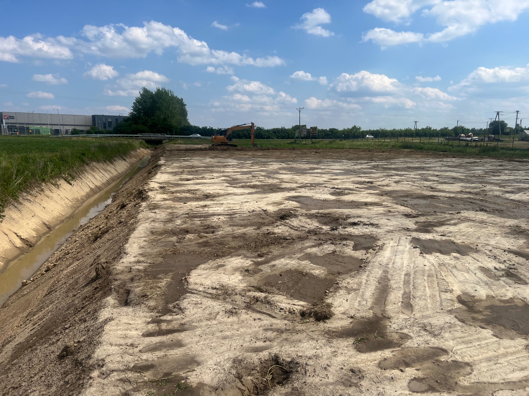 Przygotowany teren pod budowę z wykopanym rowem melioracyjnym i pracującą koparką w tle, widok w słoneczny dzień.