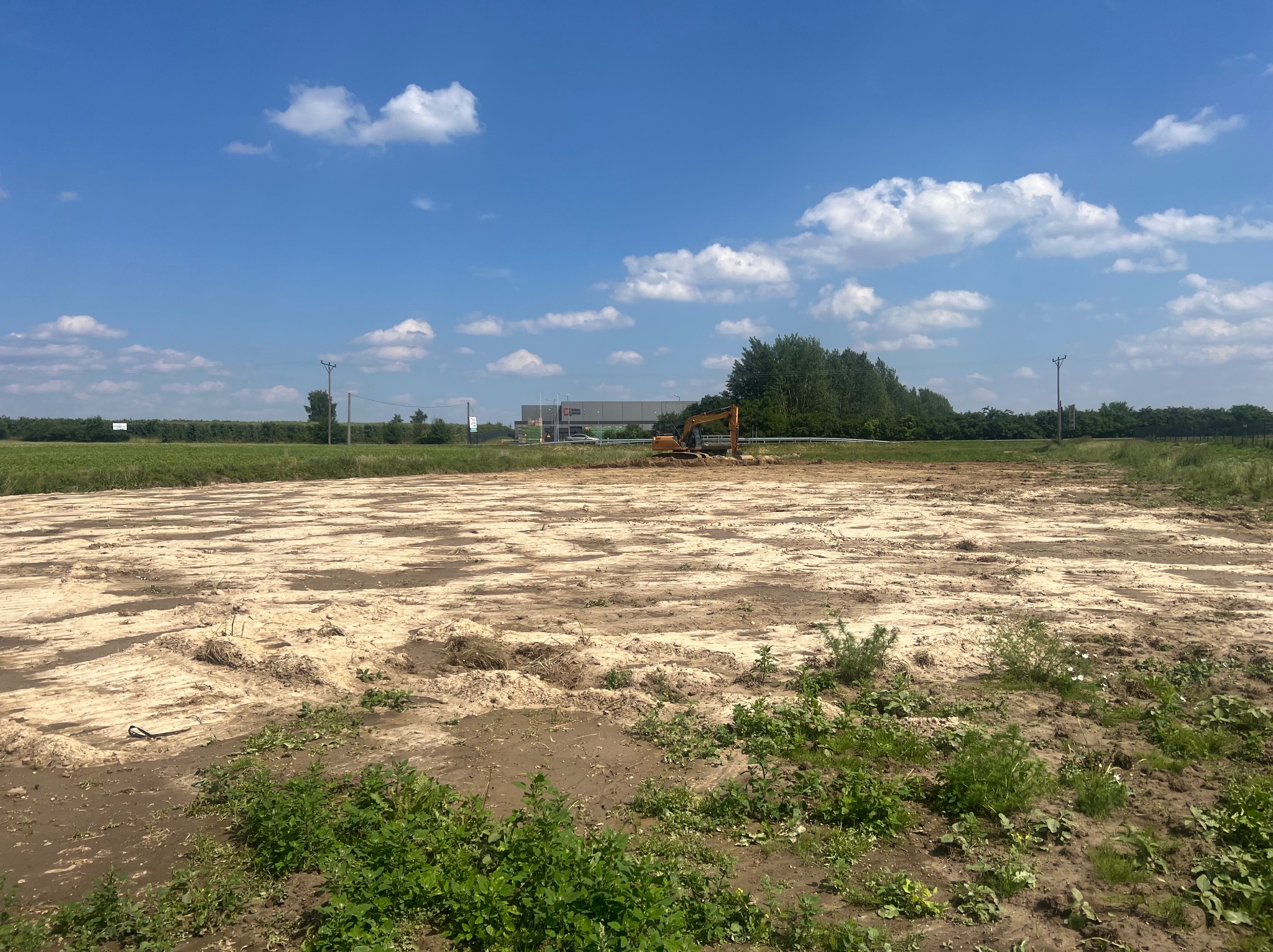Żółty, gąsienicowy koparka na wyrównanym, piaszczystym terenie pod błękitnym niebem z chmurami. W tle budynek magazynowy i zieleń. Ślady po pracy maszyny.