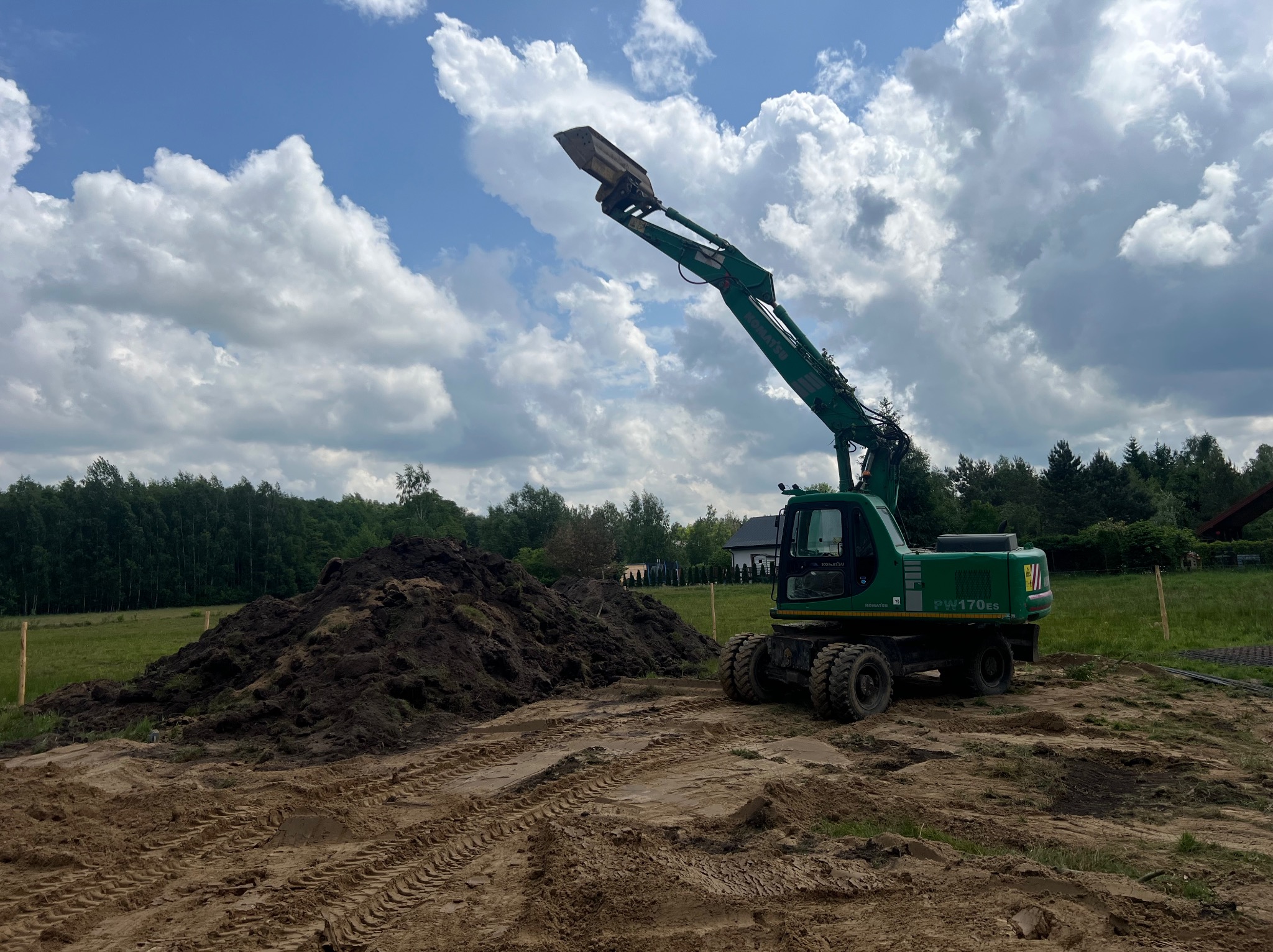 Zielona koparka Komatsu PW170ES z podniesionym ramieniem i łyżką na placu budowy, z hałdą ziemi i śladami opon w tle, pod błękitnym niebem z chmurami.
