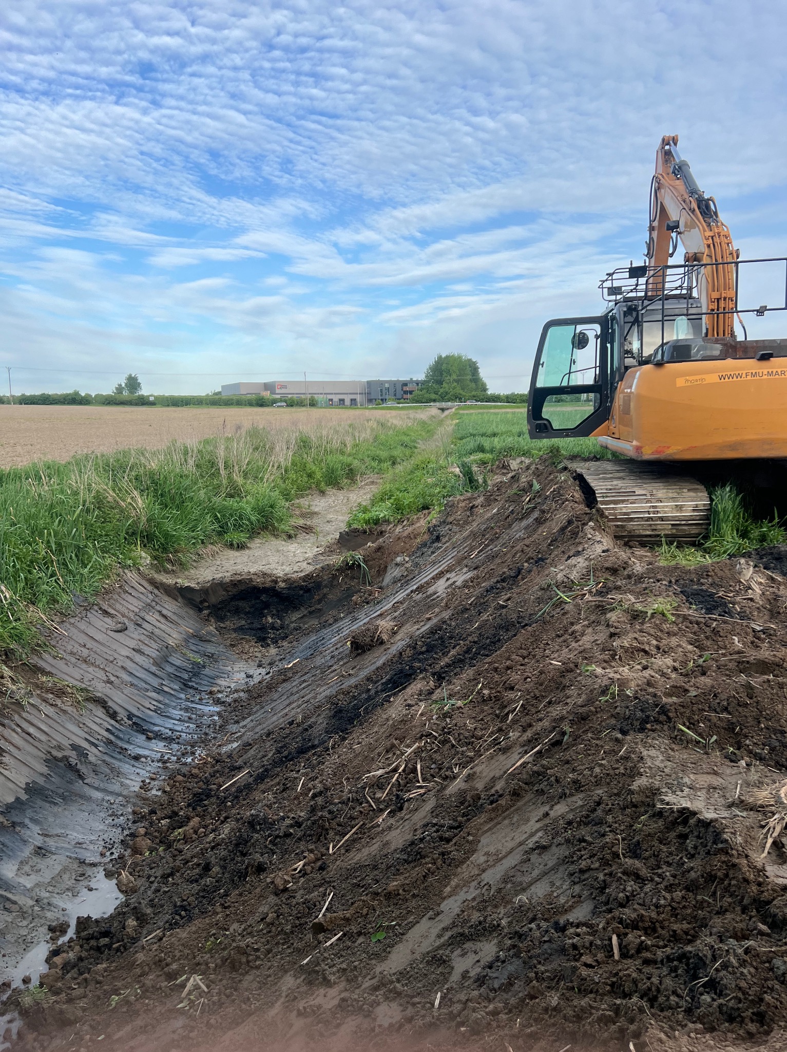 Pomarańczowa koparka gąsienicowa przy wykopanym rowie melioracyjnym, Warka. Widoczne ślady pracy maszyny i świeżo usunięta ziemia.