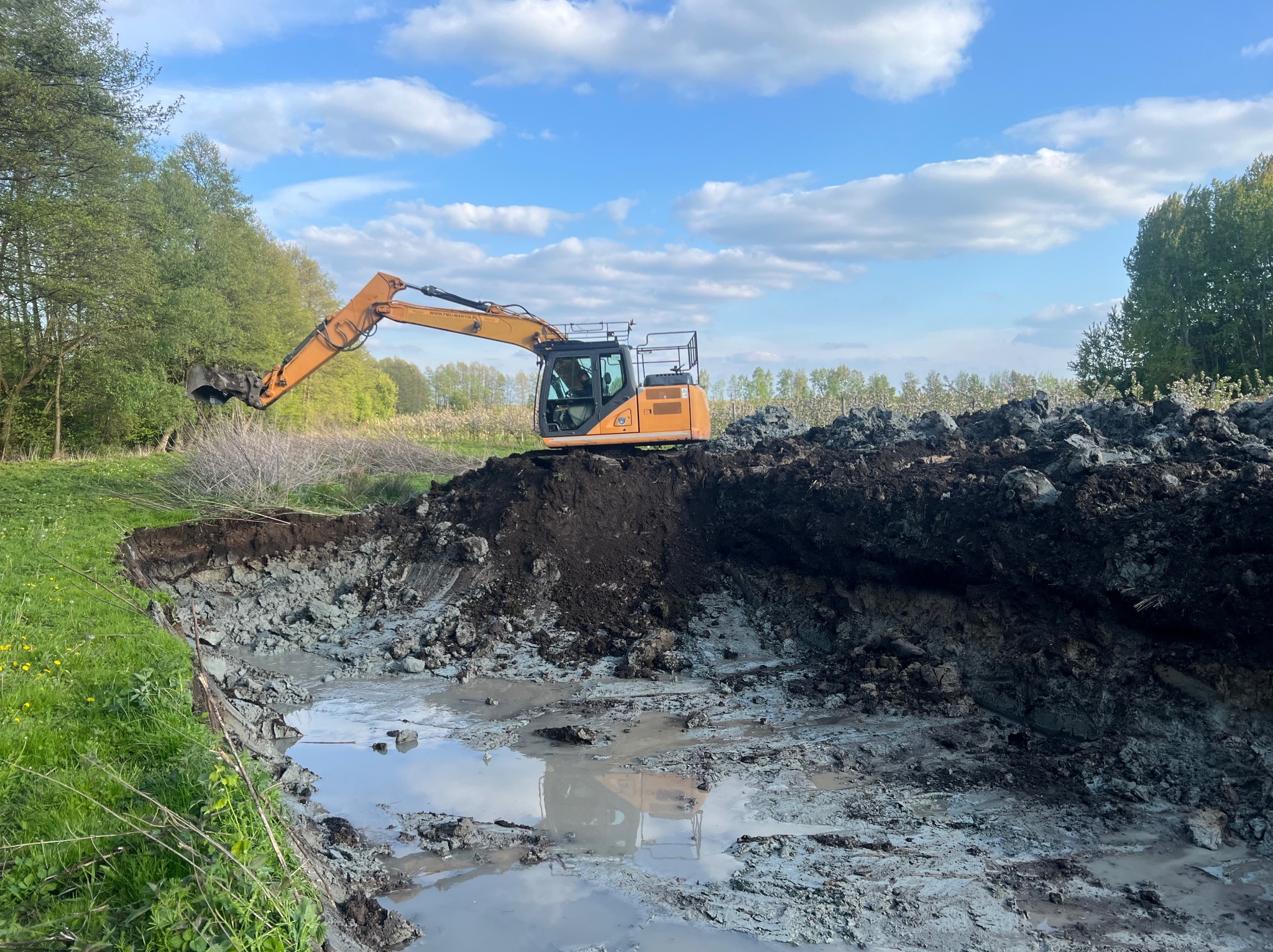 Żółta koparka na skraju wykopu pod staw, odbicie maszyny w wodzie, zielone drzewa w tle, błękitne niebo z chmurami. Wykopanie stawu.