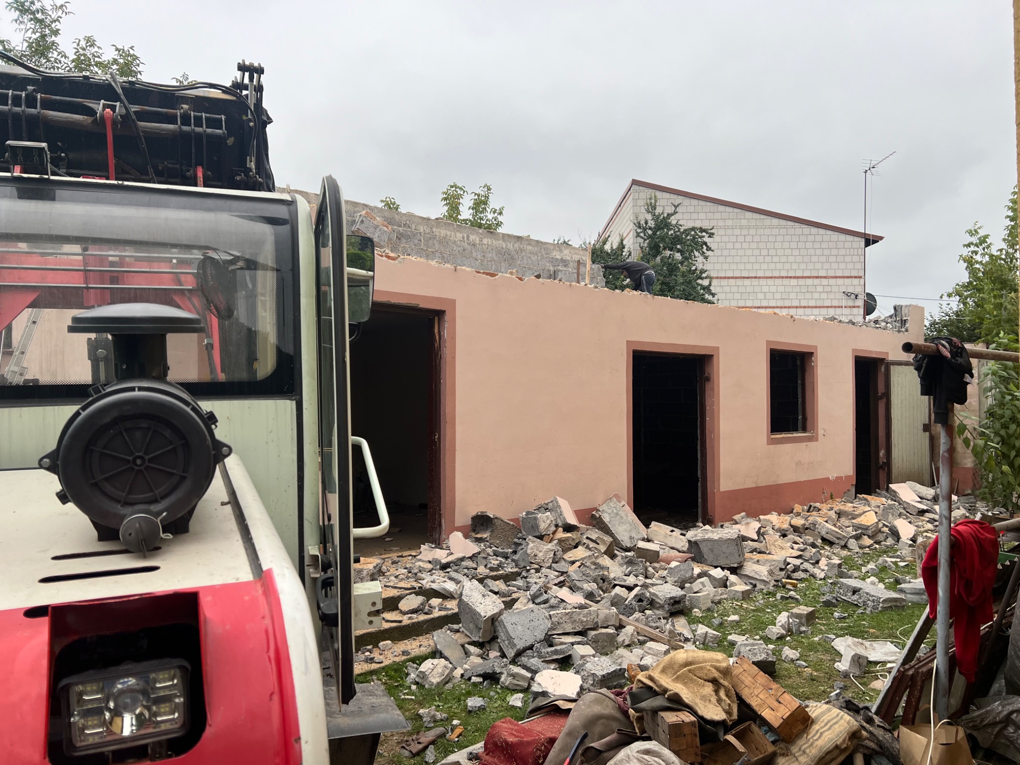 Częściowo rozebrany budynek z widoczną stertą gruzu i pracownikiem na dachu, obok zaparkowany pojazd z wysięgnikiem.