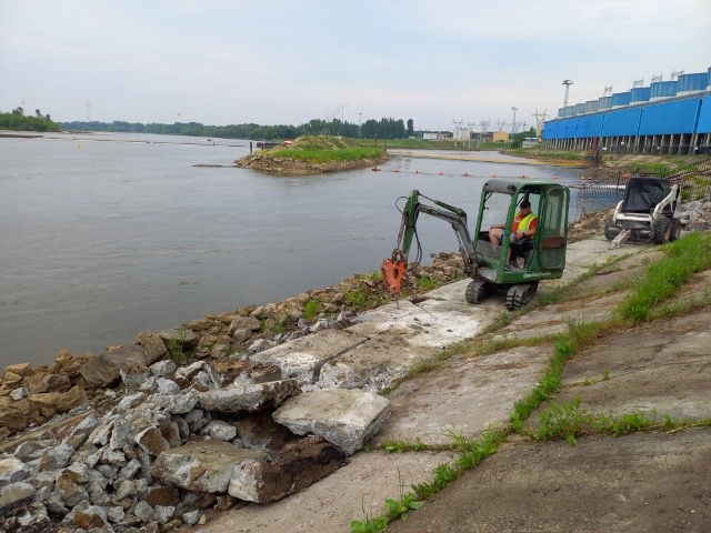 Niewielka koparka z wiertłem hydraulicznym kruszy betonowe nabrzeże rzeki; w tle widać budynki elektrowni i bobcata.