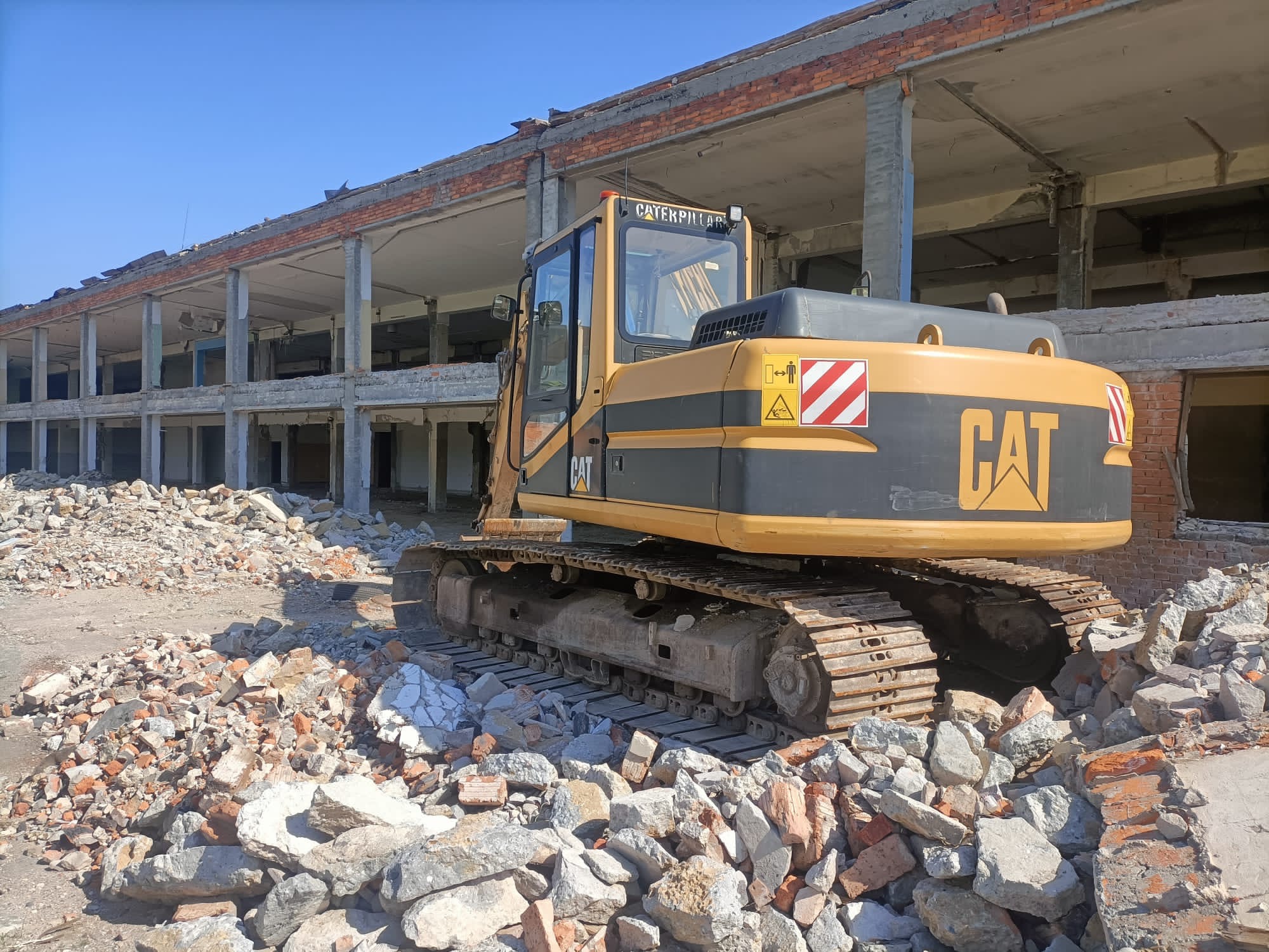 Żółta koparka gąsienicowa Caterpillar stoi na gruzowisku cegieł i betonu przed szkieletem rozbieranego budynku z betonowymi kolumnami pod błękitnym niebem.
