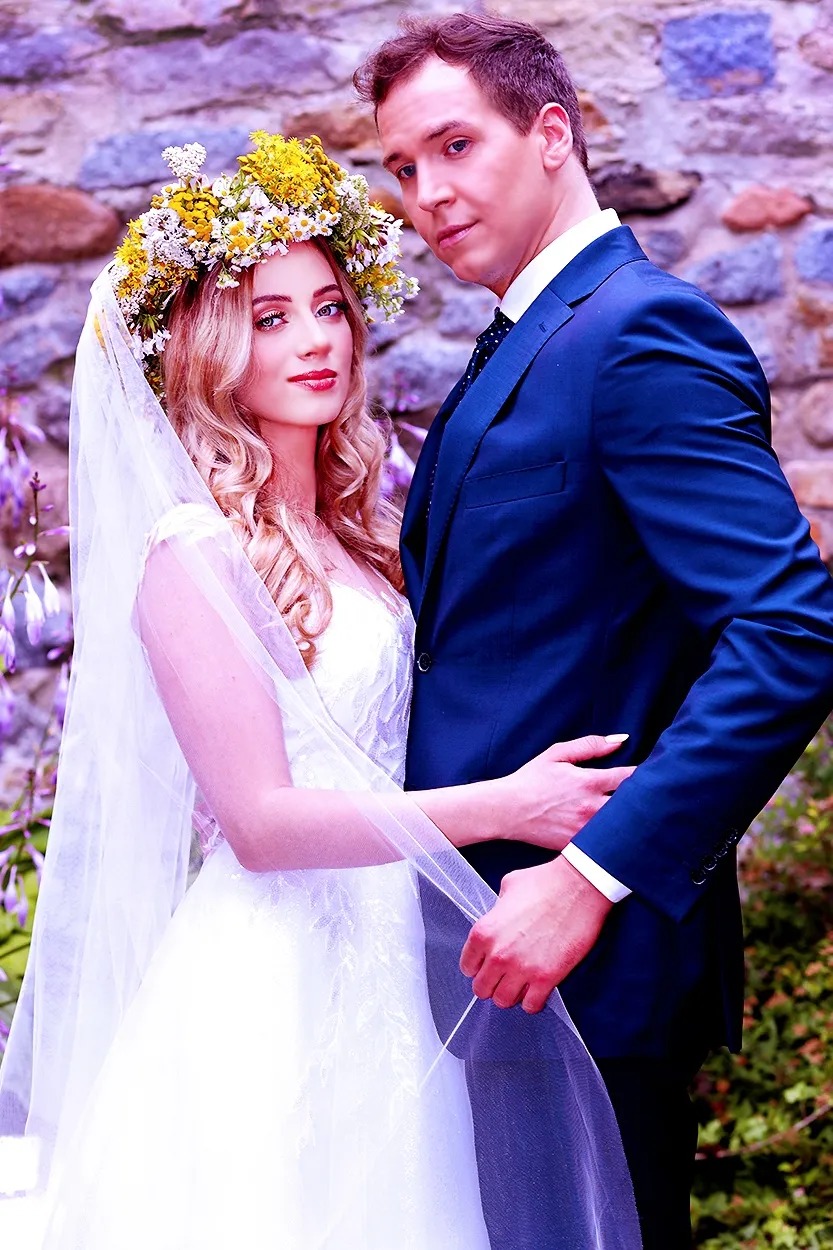 Para młoda w plenerze: panna młoda z wiankiem z żółtych kwiatów, w białej sukni i welonie, obejmowana przez pana młodego w granatowym garniturze, na tle kamiennej ściany i zieleni.