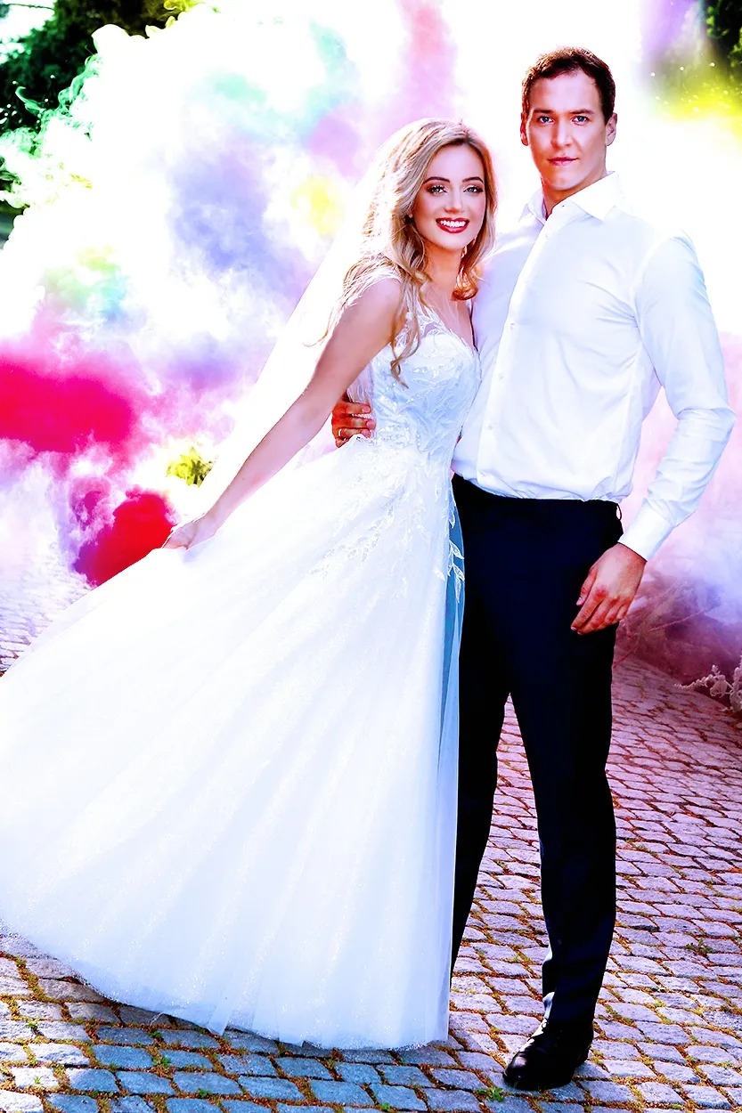 Para młoda w białej sukni ślubnej i ciemnym garniturze pozuje na tle kolorowych dymów na brukowanej drodze.