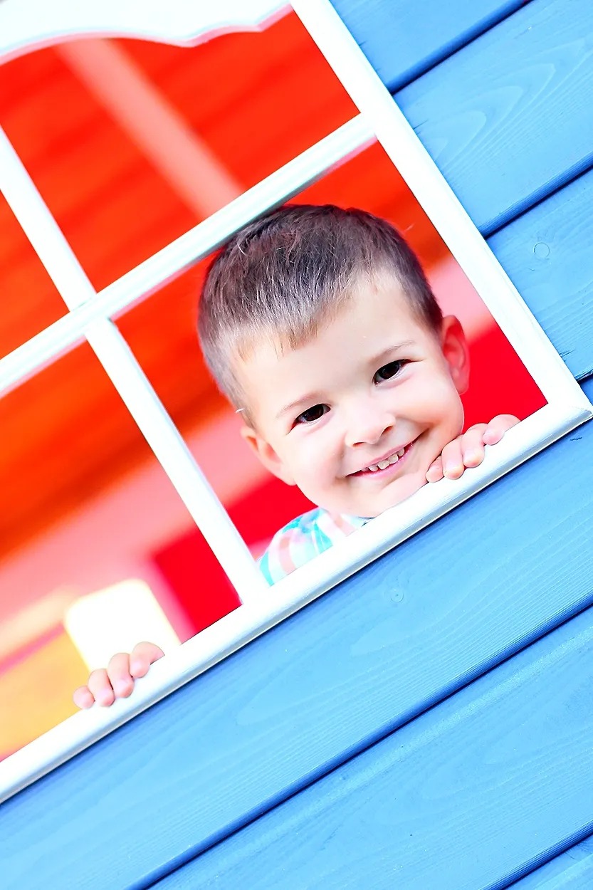 Uśmiechnięty chłopiec z brązowymi włosami wygląda przez okno niebieskiego domku zabaw z czerwonym wnętrzem.