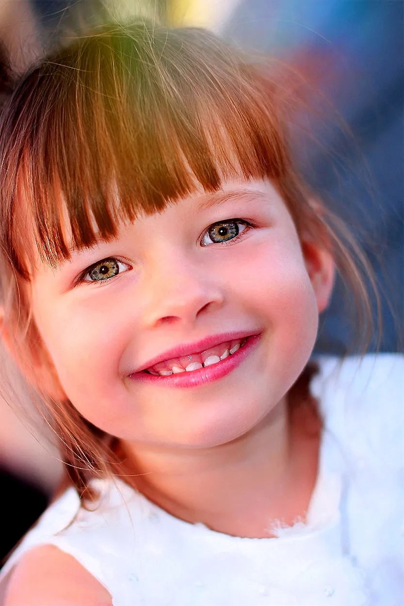 Portret uśmiechniętej dziewczynki z brązowymi włosami i zielonymi oczami, delikatny makijaż, biała bluzka.