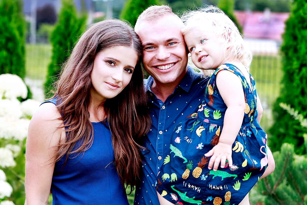 Uśmiechnięta rodzina: mama, tata trzymający córeczkę w sukience z motywem tropikalnym, na tle zielonych krzewów.