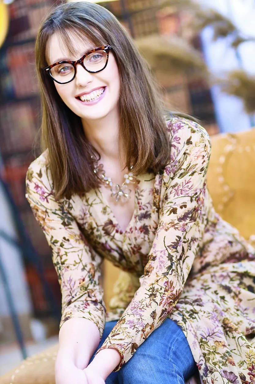 Portret uśmiechniętej kobiety w okularach w rogowej oprawce, ubrana w kwiecistą sukienkę i dżinsy, siedzącej na fotelu na tle regału z książkami.