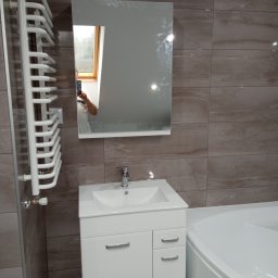 Rafał Jędrzejczyk - Nowoczesna łazienka na poddaszu z brązowymi płytkami imitującymi marmur, białą szafką pod umywalkę, grzejnikiem drabinkowym i narożną wanną; w lustrze odbija się okno dachowe i osoba robiąca zdjęcie.