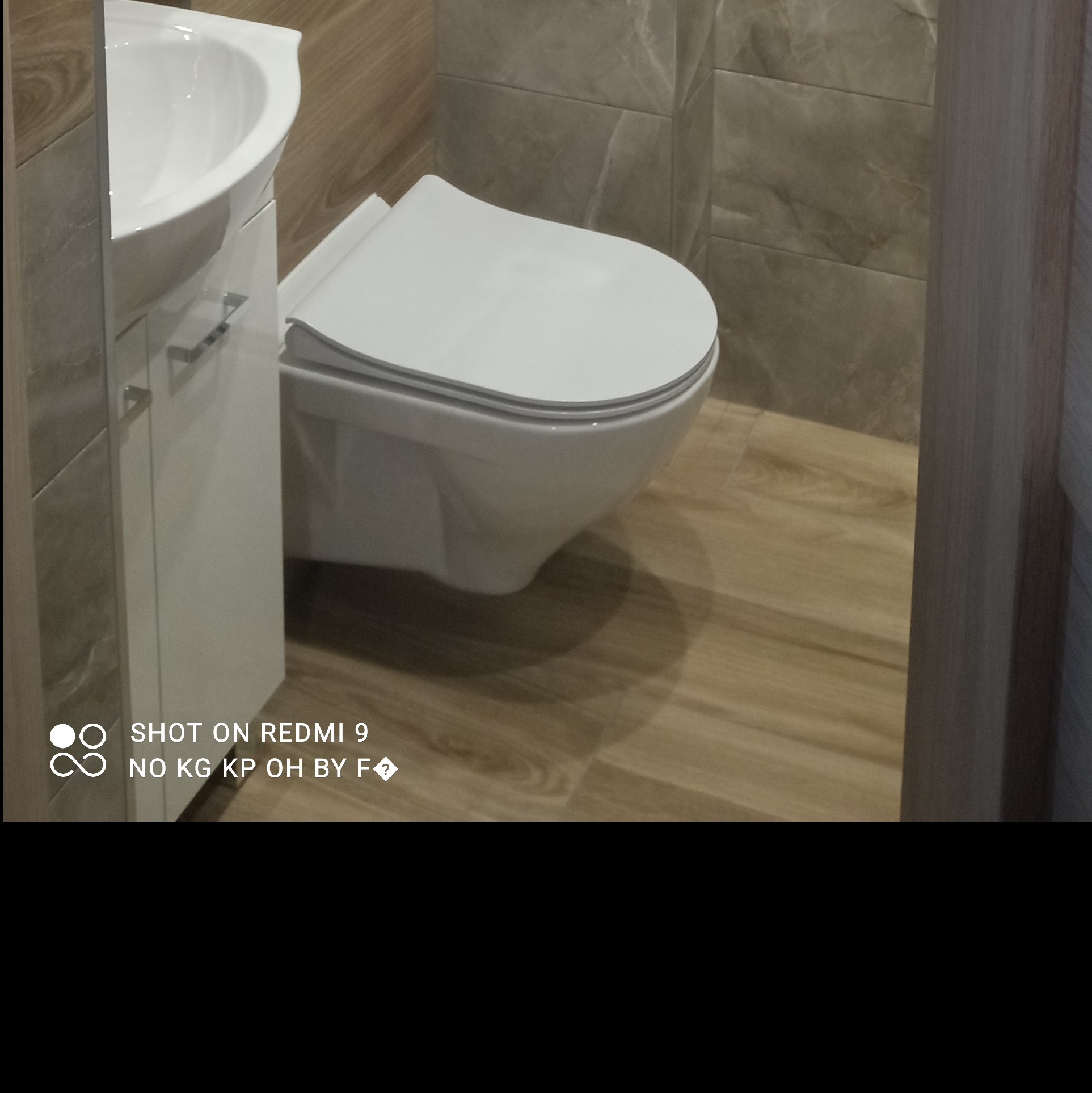 Wisząca toaleta z deską wolnoopadającą i umywalka z szafką w nowocześnie wykończonej łazience z płytkami imitującymi drewno i kamień.