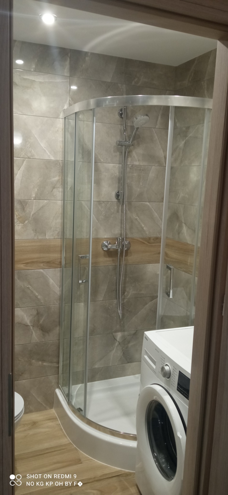 Kabina prysznicowa z chromowanymi elementami i deszczownicą, obok pralka w wykończonej łazience z płytkami imitującymi marmur i drewno.