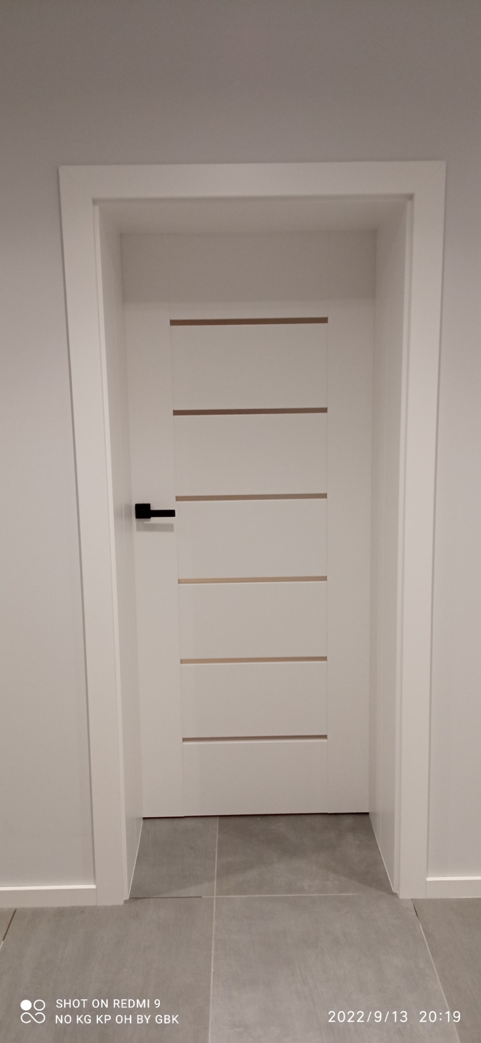 Zamknięte, białe drzwi wewnętrzne z poziomymi, drewnianymi wstawkami i czarną klamką, osadzone w białej ościeżnicy, na tle szarej ściany i podłogi z płytek.