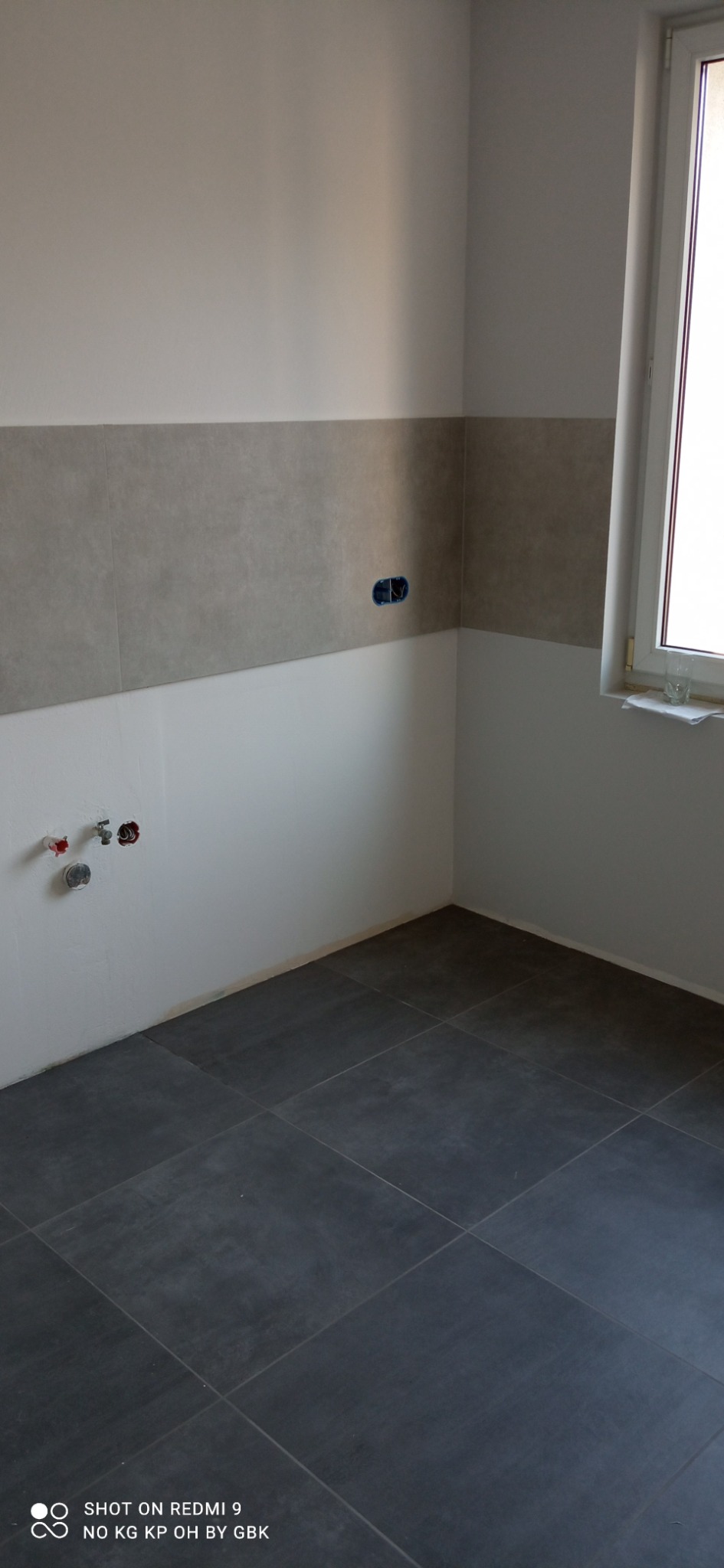 Fragment łazienki w trakcie prac wykończeniowych: szare płytki na podłodze, częściowo położone szare płytki na ścianie, widoczne wyprowadzenia instalacji wodnej i gniazdko elektryczne.