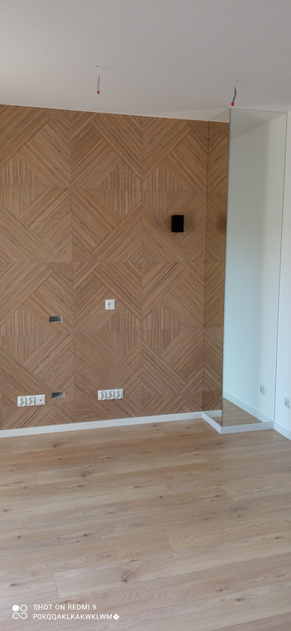 Ściana z dekoracyjną, drewnianą okładziną w geometryczne wzory, z widocznymi gniazdkami elektrycznymi i otworami na kable, oraz fragmentem lustra, na podłodze panele w odcieniach beżu.