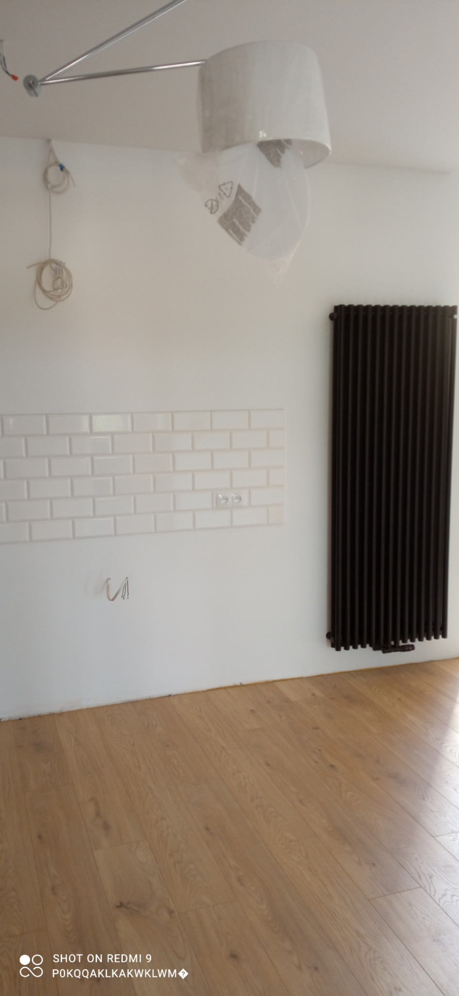 Fragment wnętrza w trakcie prac wykończeniowych: ułożone białe płytki imitujące cegłę nad gniazdkami elektrycznymi, czarny grzejnik żeberkowy, drewniana podłoga, lampa wisząca w folii ochronnej...