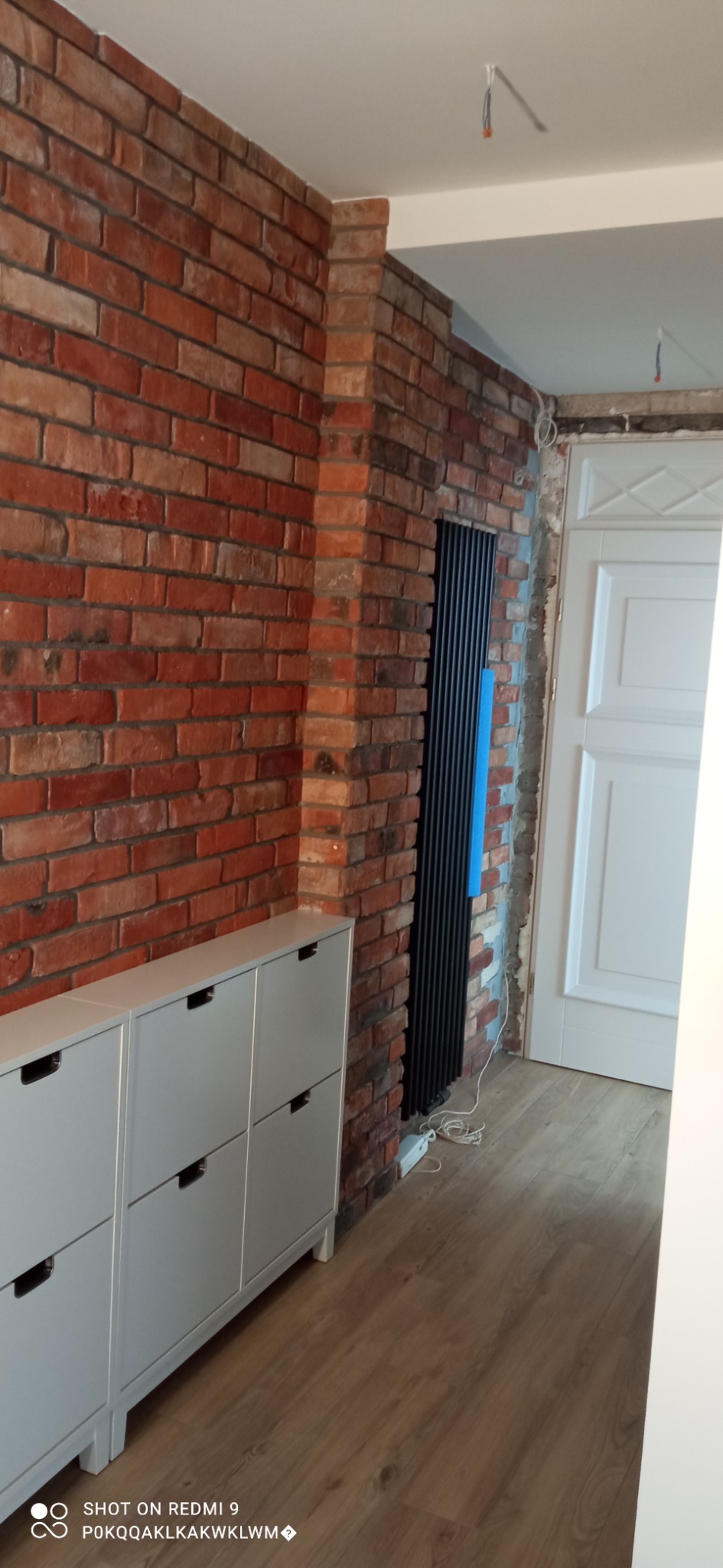 Ceglana ściana w korytarzu, z szarą szafką na buty i czarnym grzejnikiem panelowym, częściowo wykończona, widoczne przewody elektryczne.